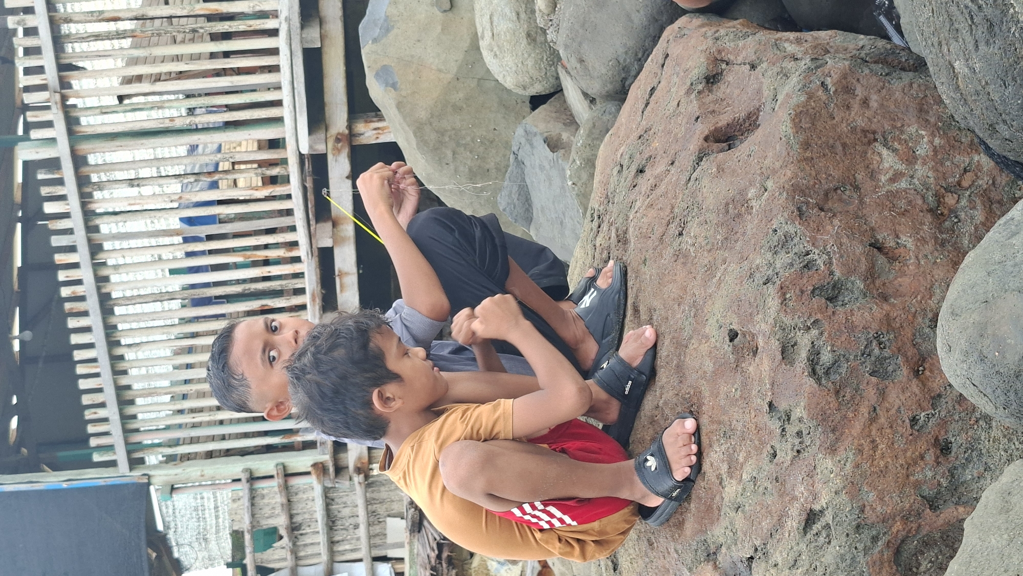 Two young boys are fishing on a rocky shore in Lhokseumawe, Indonesia. The boy in the foreground is wearing a mustard-colored t-shirt, red shorts with white stripes, and black Adidas slides. He is crouching on a large, reddish-brown rock, concentrating on the fishing line he is holding. To his left and slightly behind, another boy in a gray shirt and black pants is also crouching, looking towards the water. In the background, a wooden structure with a fence is visible, suggesting the area is near a boardwalk or a pier. The overall environment is natural, with a focus on the activity of fishing.