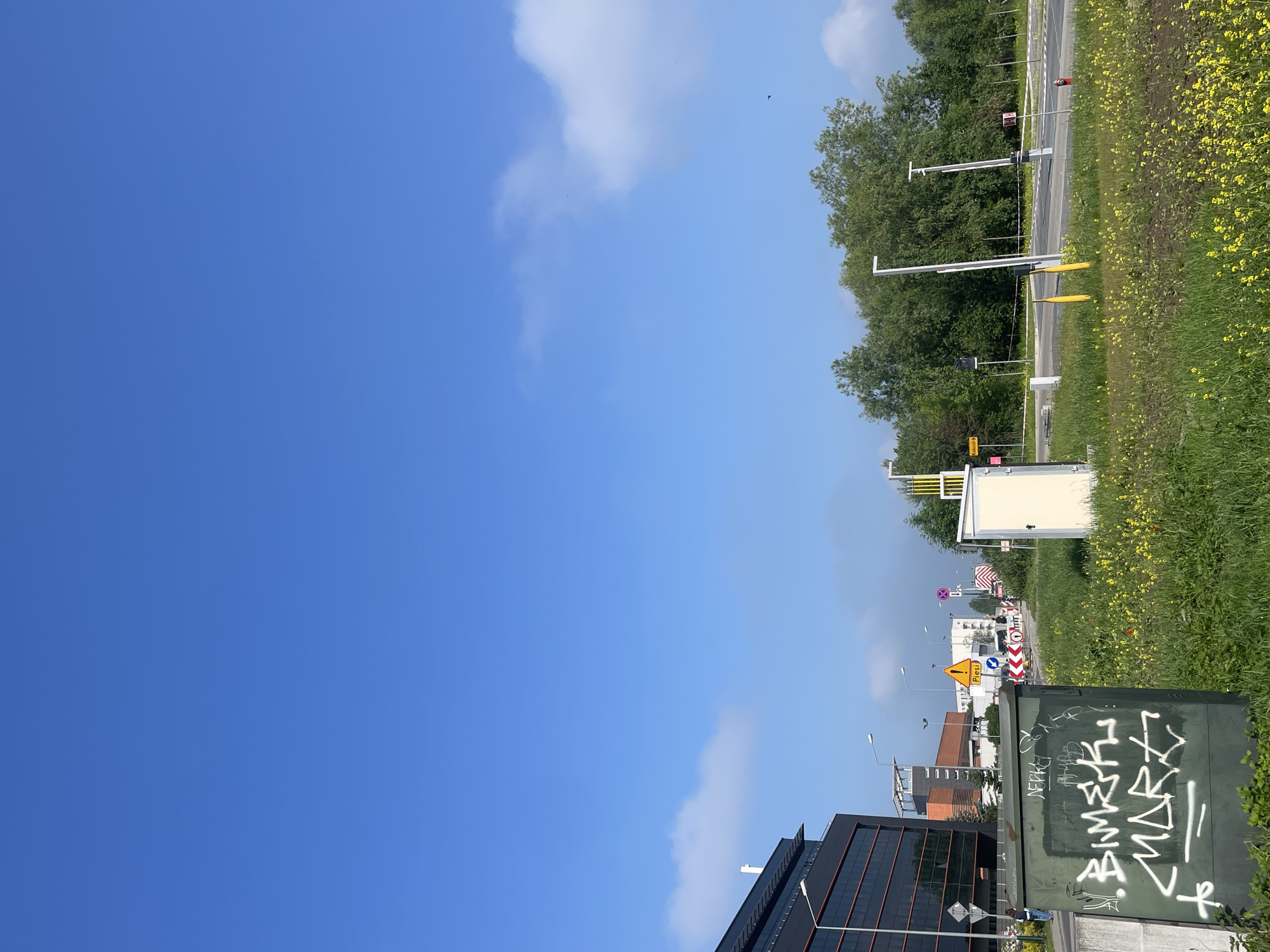 The image depicts an urban or suburban outdoor scene, dominated by a clear blue sky with scattered white clouds. The perspective appears to be from ground level, looking towards a road and buildings.
In the foreground, a large, dark green utility box, heavily covered in white graffiti and stylized symbols, occupies the bottom right corner. To its left, partially visible in the bottom left, is a modern building featuring dark glass windows and a flat roof.
Mid-ground elements include a road or highway running vertically along the right side of the frame. This road is bordered by a wide grassy verge densely populated with small yellow wildflowers. A line of green trees forms a natural barrier behind the road. Along the road, various infrastructure elements are visible, such as tall poles for streetlights and traffic signals. Several road signs are present, including a yellow triangular warning sign with a black exclamation mark, and a white rectangular sign with black text that clearly reads "Piesi" (Pedestrians in Polish) and "przejście" (crossing in Polish). Other signs, like a red circular prohibitory sign, are also visible but less clear. Further down the road, more buildings are visible in the distance.
The scene is brightly lit, indicating it is daytime, likely morning or afternoon, under clear weather conditions with some light cloud cover. Small, dark specks, possibly birds or insects, are faintly visible against the bright sky. No human activity is overtly depicted, suggesting a static street view.