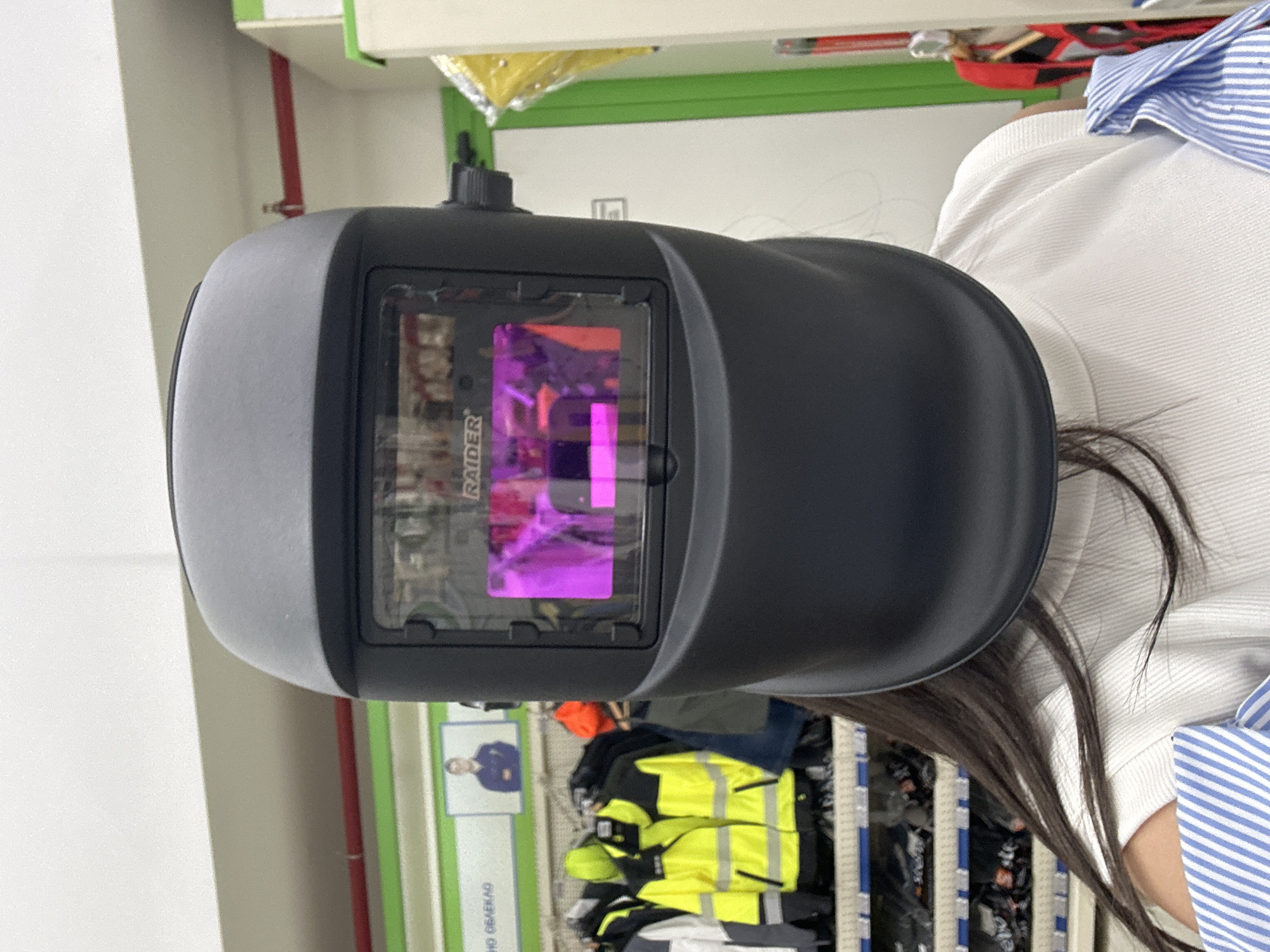 A person is wearing a black welding helmet in an indoor retail environment. The helmet is the primary subject, covering the person's face and upper head. Through the helmet's visor, a distorted, purple-hued reflection or display is visible, possibly indicating the helmet's functionality. The person is wearing a white ribbed top and a blue and white striped garment on their left arm. In the background, shelves stocked with various items, including high-visibility jackets and other workwear, are visible. A poster with an image of a person in a dark blue shirt is also present on the wall. The environment appears to be a store selling safety equipment or work clothing.