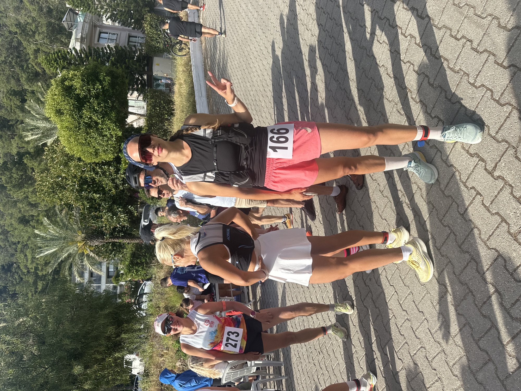 The image captures a group of female runners, likely participants in a race or event, in Fethiye, Turkey. The setting appears to be a paved area, possibly a street or sidewalk, with a building and lush greenery in the background, suggesting an urban or suburban environment. The runners are wearing athletic attire with visible race numbers: 273, a runner in the foreground, 166. The runners have a range of styles and colors, and the presence of race numbers indicates that this may be a competitive event. The runner with the number 166 is flashing a peace sign. The lighting suggests it is a sunny day. Other people are in the background.
