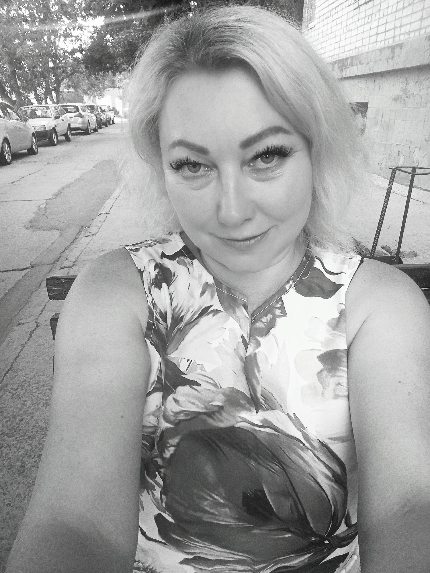 A black and white, eye-level, selfie style, medium shot shows a middle-aged woman with blonde hair and prominent eyelashes. She is sitting on a bench outdoors, with a street lined with parked cars visible behind her. The woman is wearing a floral print sleeveless top. The environment appears to be a city street with trees and a brick building in the background. There is a metal grate or railing to the right of the woman. The overall scene is an outdoor, urban environment.