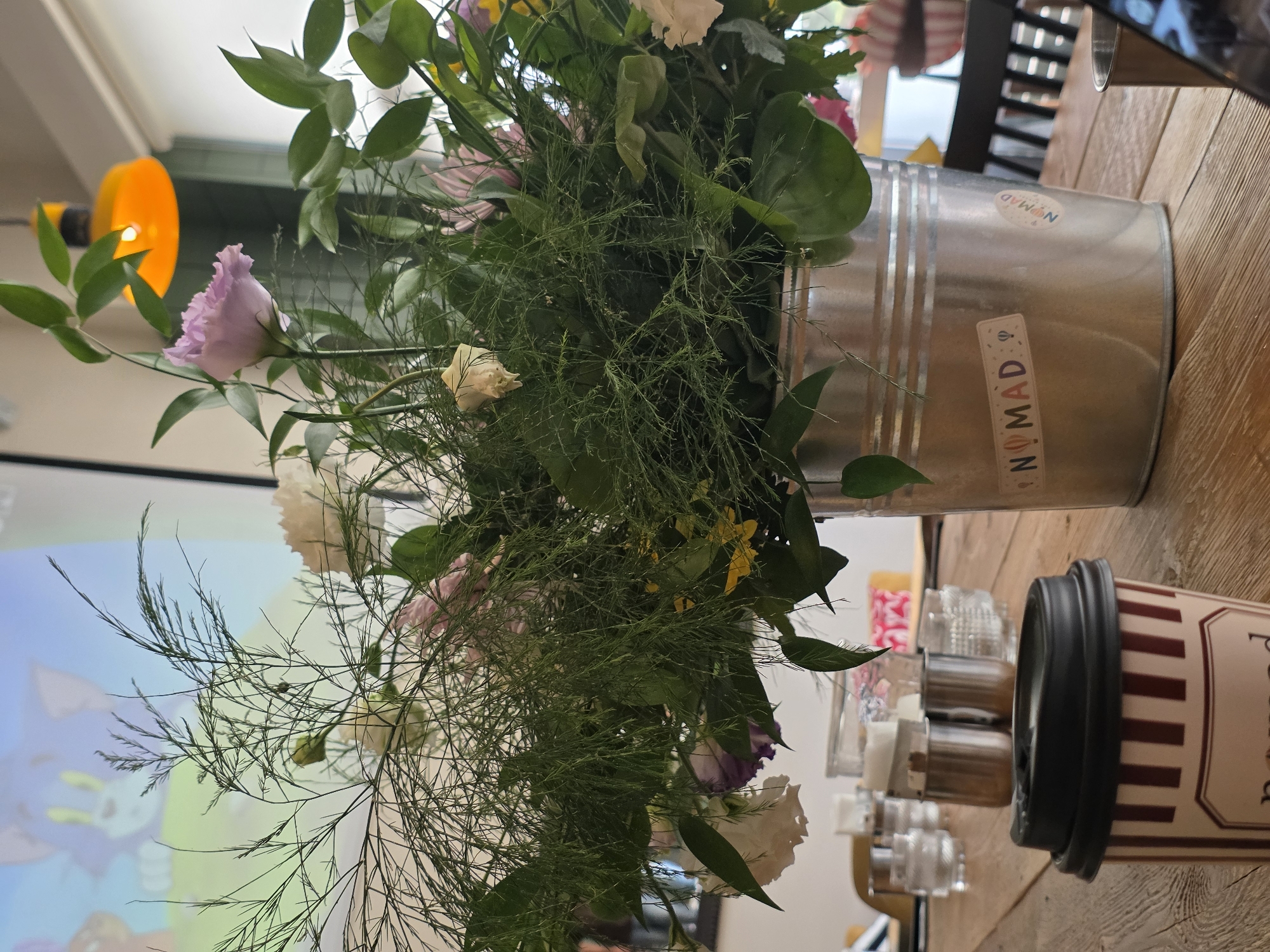 A close-up shot captures a bouquet of flowers in a galvanized metal bucket placed on a wooden table. The bouquet is a mix of green foliage, delicate light purple and white flowers, and some yellow accents. Two stickers with the word "NOMAD" and balloon graphics are affixed to the bucket. In the foreground, a takeaway coffee cup with a black lid and a striped pattern of maroon and cream is visible. To the left of the bucket, several glasses and metal containers are arranged on the table, suggesting a dining or cafe setting. In the background, a projection screen displays an image of cartoon characters, possibly from "Tom and Jerry." An orange light fixture hangs from the ceiling. The scene appears to be indoors, with a soft, ambient light. The location is not specified within the image itself.