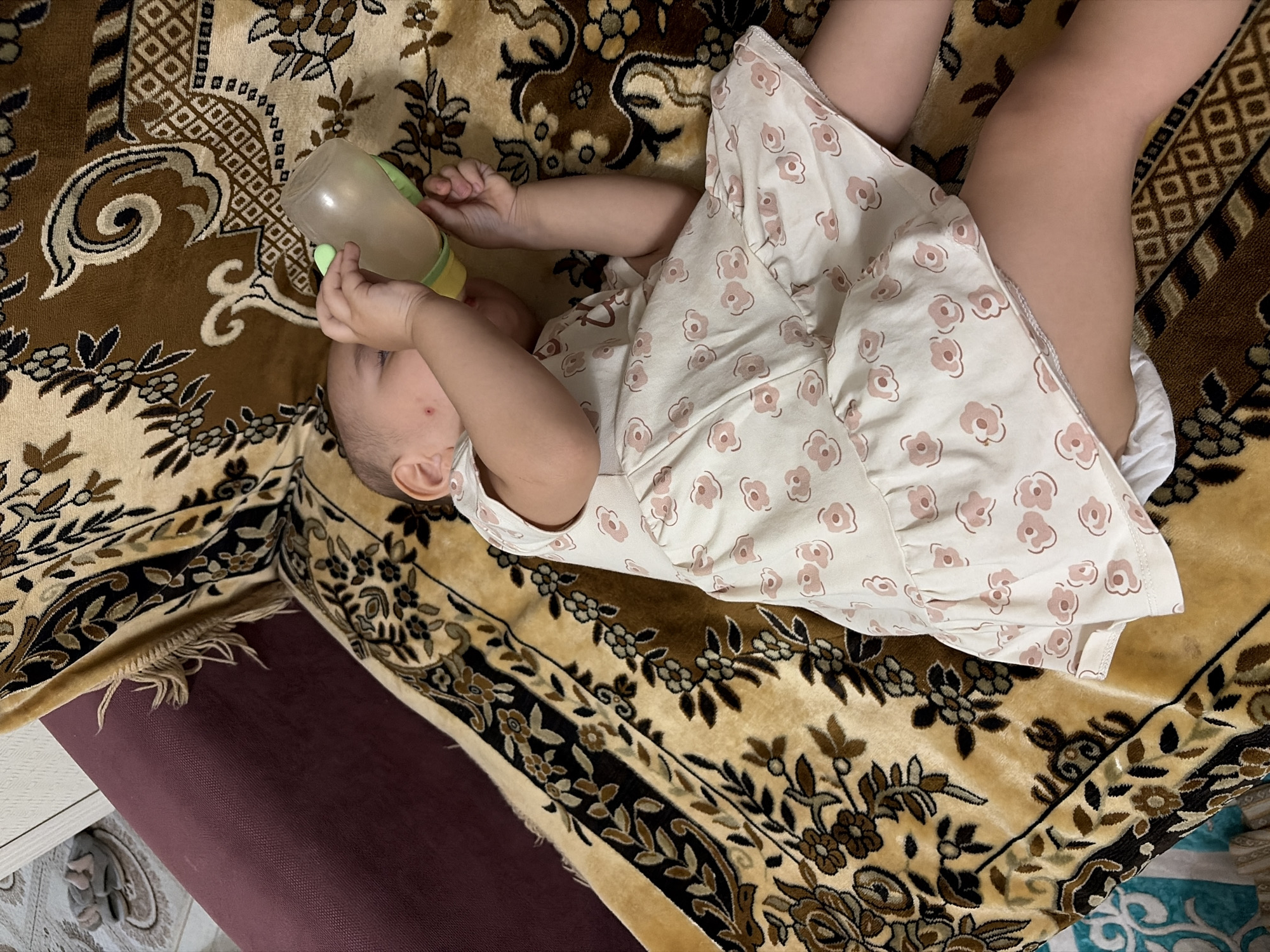 The media file shows a close-up, high-angle view of a baby lying on a patterned rug, drinking from a bottle. The baby is wearing a light-colored dress with a floral pattern. Their hands are holding the bottle, which appears to be made of plastic with a green nipple. The baby's face is partially visible, showing their ear and a small red mark on their cheek. The rug has a traditional, ornate design with shades of brown, beige, and black, featuring floral motifs and geometric patterns. In the background, a portion of a dark red surface is visible, possibly a piece of furniture. The lighting suggests it is daytime. The scene conveys a sense of domestic comfort and care. There is no visible text in the image. The location context provided is Astana, Kazakhstan.