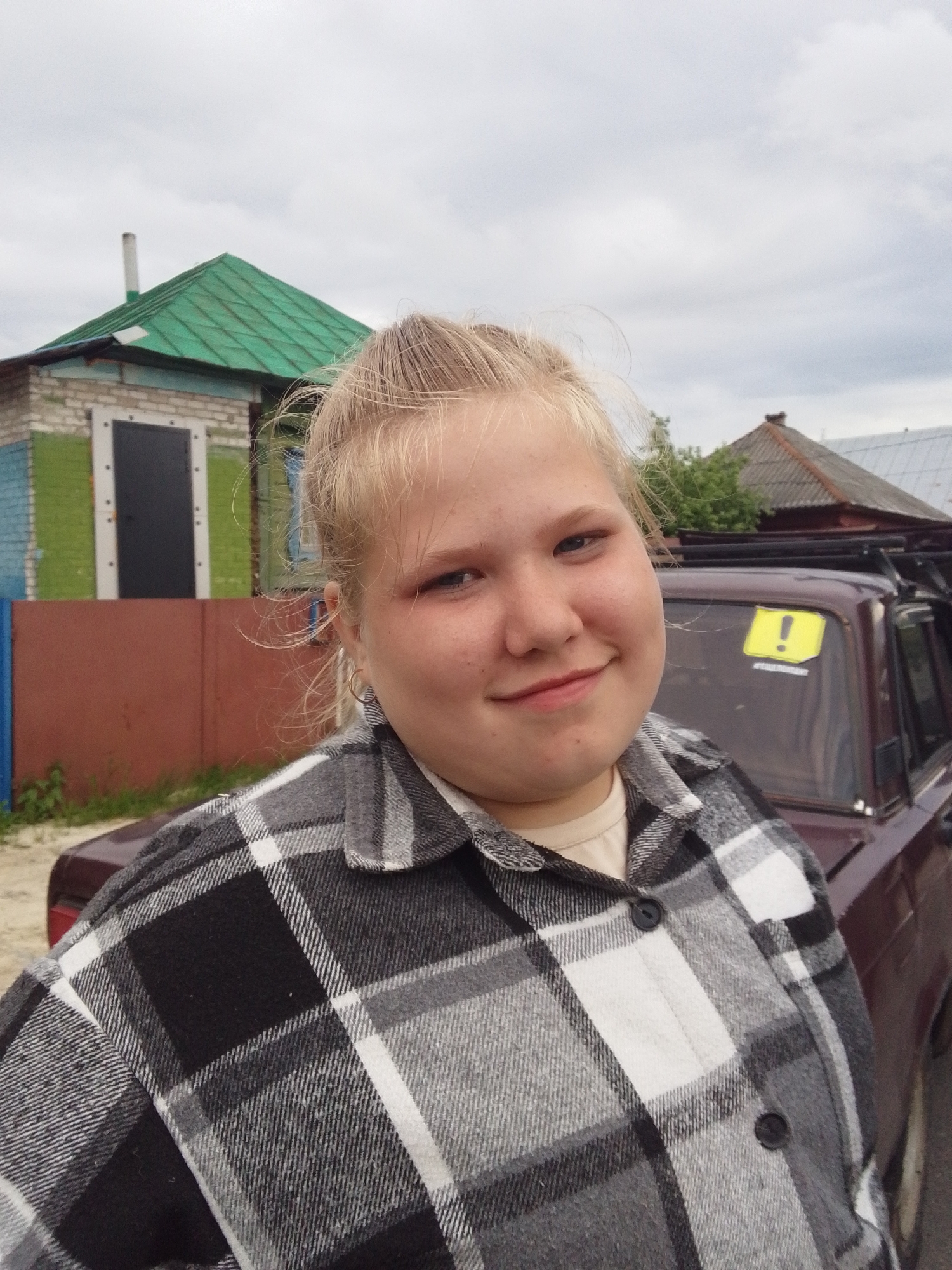 A fair-skinned, young woman with blonde hair stands in front of a burgundy car with a roof rack. She wears a gray and black plaid shirt. Behind her are a green-roofed building, a rust-colored fence, and other houses, suggesting a residential area. A yellow sticker with an exclamation point is affixed to the car's back window. The sky is overcast. The location appears to be in an urban setting.