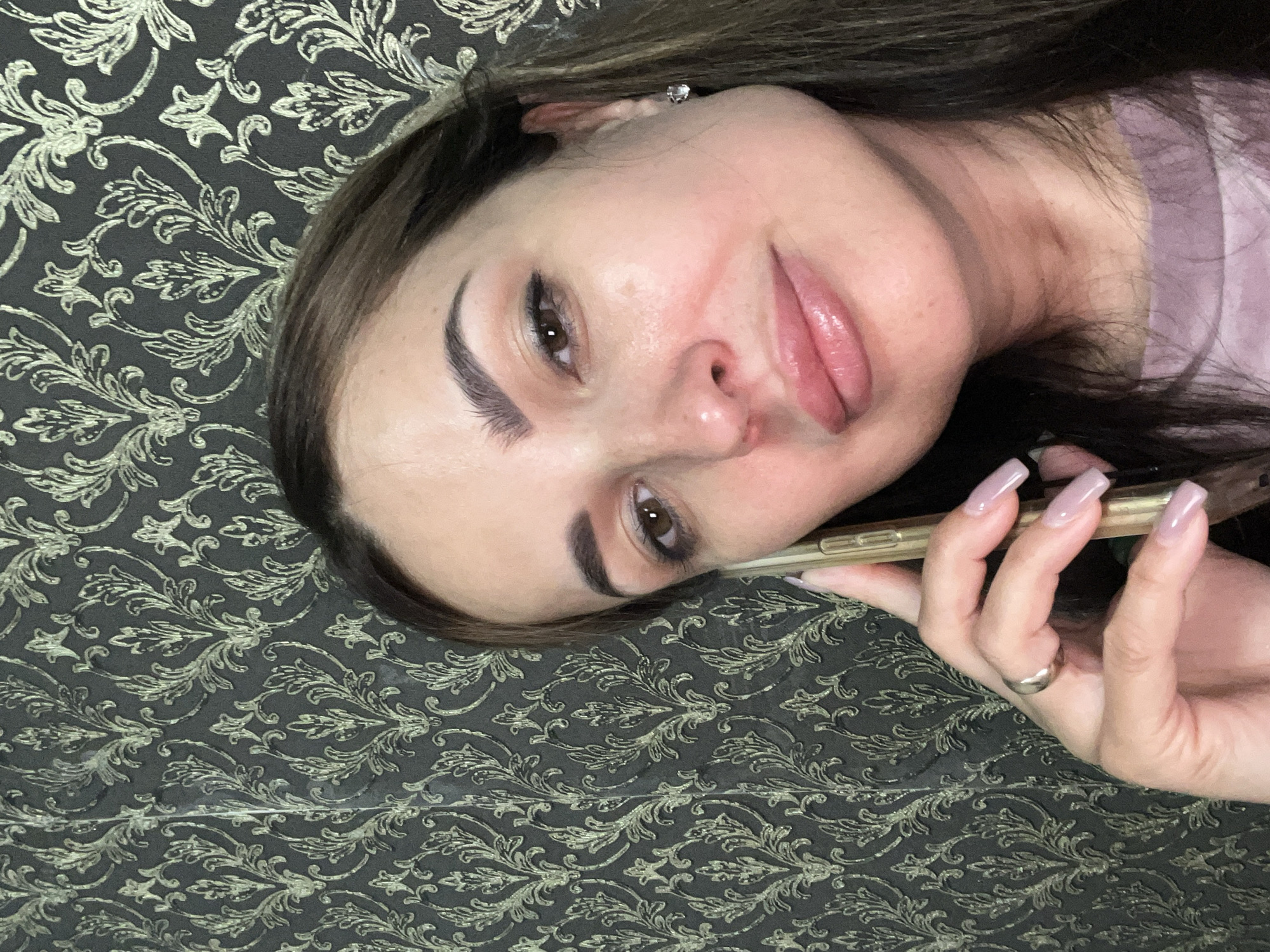 A medium shot shows a woman in her late 20s or early 30s speaking on a smartphone, held to her right ear and cheek. She is positioned in the center right of the frame, facing the viewer with her head tilted slightly to the left. The woman has long, dark brown hair that falls around her shoulders and is wearing a light purple t-shirt. Her makeup includes eyeshadow, eyeliner, and lip gloss, and she has a diamond stud earring in her right ear. A silver ring is visible on her left ring finger. The background is a dark grey wallpaper with a repeating gold damask pattern.