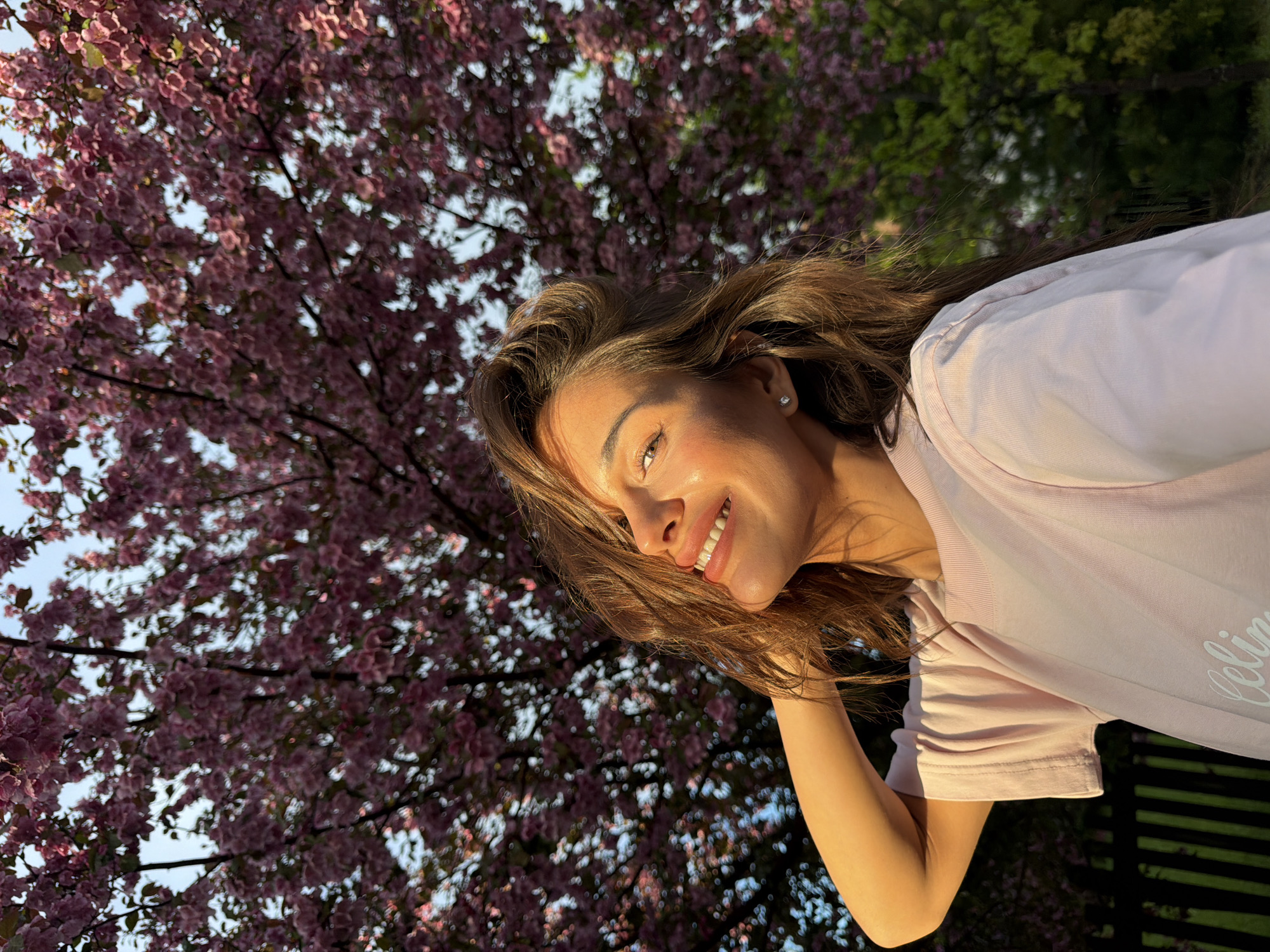A woman smiles while posing against a backdrop of pink-blossomed trees. She has brown hair and is wearing a light pink t-shirt. A partial view of a dark fence can be seen in the lower right corner. The location in Shamoni, Russia cannot be determined based on the given image.