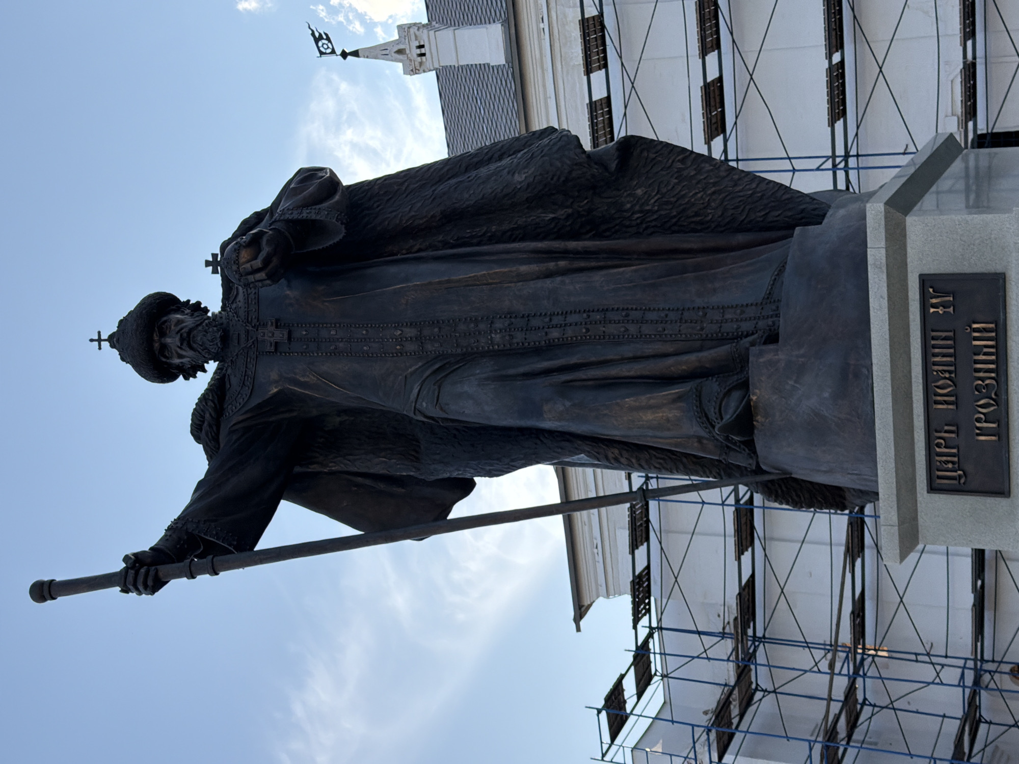 A bronze statue of Ivan the Terrible stands in the city of Vologda, Russia, in front of a white building with a grey roof. The statue depicts Ivan the Terrible holding a scepter in his raised left hand and an orb in his right. He wears a fur-lined robe, a tall, fur-trimmed hat with a cross on top, and a beard. The statue is on a stone pedestal with a bronze plaque that reads "ЦАРЬ ИОАНН IV ГРОЗНЫЙ" (Tsar Ivan IV the Terrible). Blue scaffolding is set up around the building behind the statue, and a white tower with a dark flag is visible on the right side of the building. The sky is blue with some white clouds.
