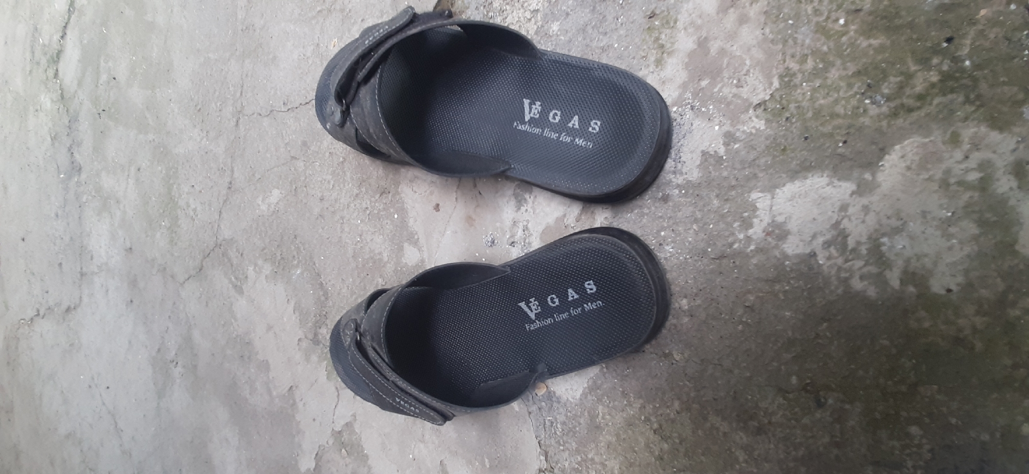 The image shows a pair of dark grey VEGAS flip-flops with the text "Fashion line for Men" printed on the soles. The flip-flops are lying on a rough, concrete surface. The concrete appears weathered and has some cracks and discolored patches, suggesting it might be an outdoor setting. The lighting is natural, indicating it could be daytime. The scene is static, with no activity taking place beyond the placement of the flip-flops. No people are visible in the image. The background is an out-of-focus, mottled grey and green concrete ground. The image appears to be taken from a slightly elevated perspective, looking down at the flip-flops.