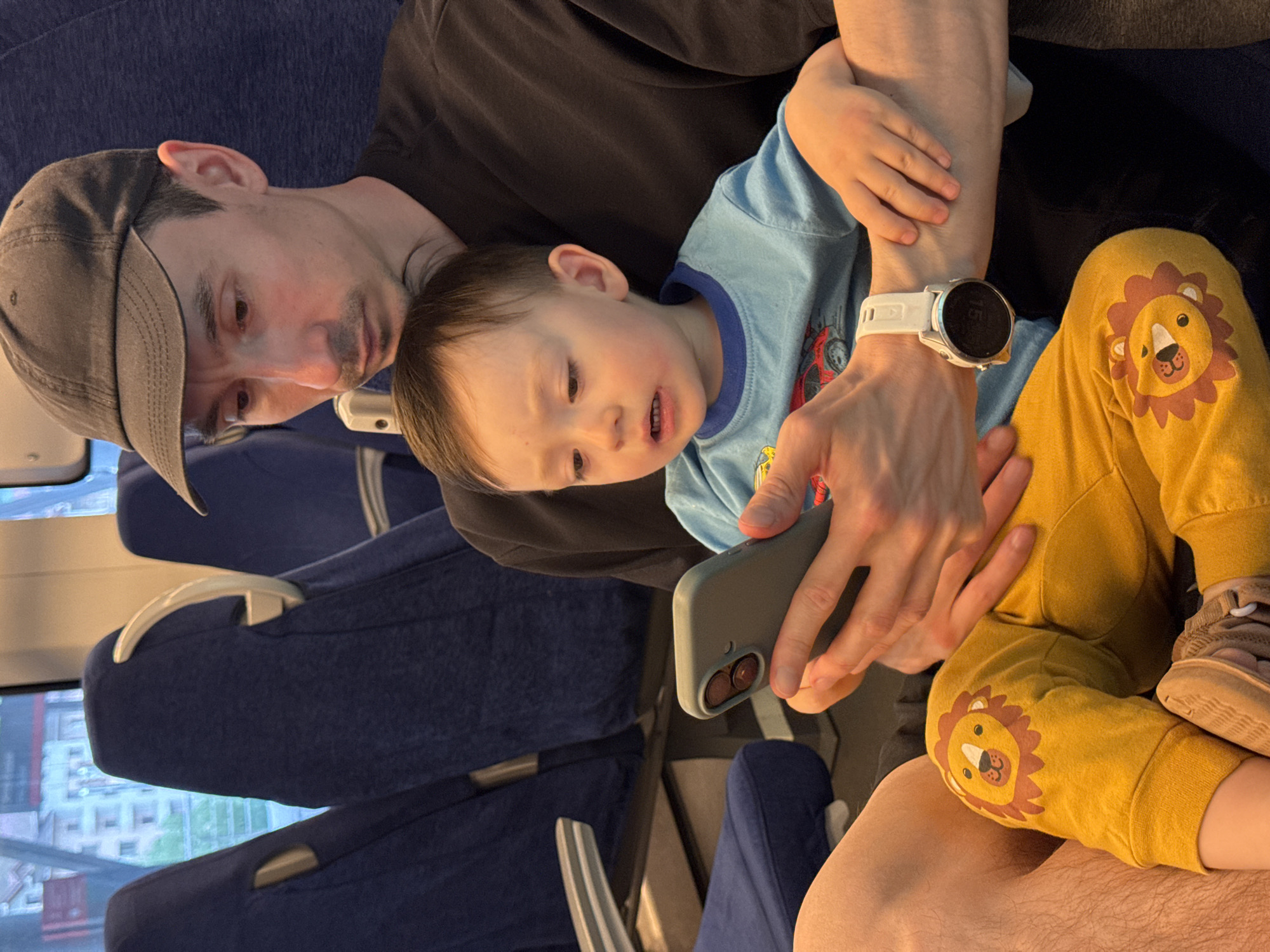 A man wearing a dark baseball cap, a dark gray t-shirt, and a white smartwatch is seated on a train with a young child wearing a blue t-shirt and yellow pants with lion designs on the knees. The child is nestled in the man's arms and is looking at a mobile phone being held by the man. The train seats are blue, and the scenery visible through the window appears to be an urban landscape with buildings and greenery, indicative of the city of Estosadok, Russia.