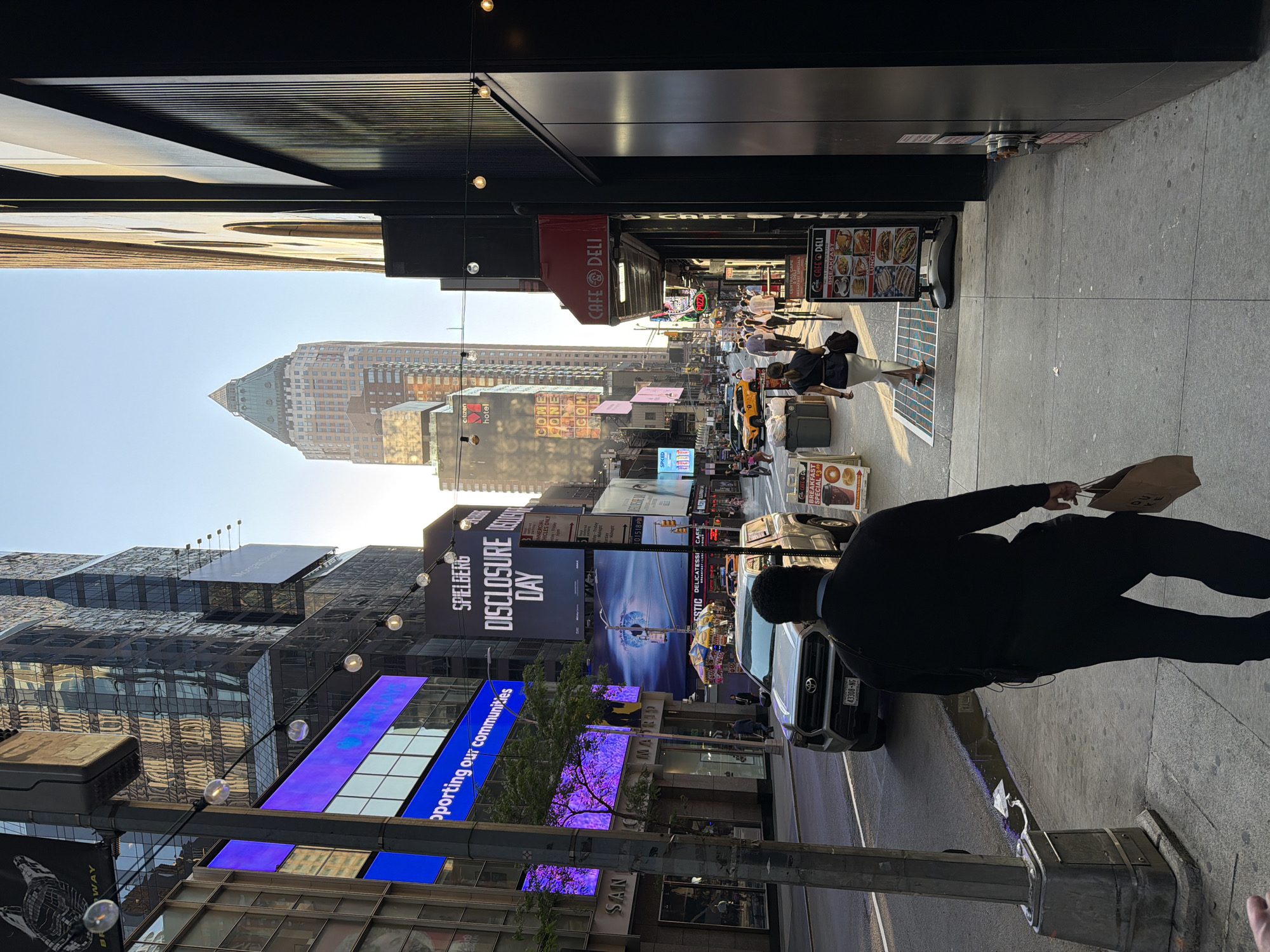 Here is a description of the image:
The image captures a bustling city street, likely in New York City, indicated by the towering buildings and vibrant advertisements. The shot is taken at street level, with a pedestrian walking in the foreground on the right side of the frame. The pedestrian is wearing dark clothes. A parked Toyota Tacoma is next to the pedestrian. Further down the street, there is a "CAFE DELI" and people milling about. Numerous large digital billboards display various advertisements and messages, including promotions for "SPIELBERG DISCLOSURE DAY" and a message about "supporting our communities". Buildings of varying heights line the street, reflecting the sunlight and adding to the urban landscape. The sky is visible between the buildings, suggesting that the photo was taken in the late afternoon as a warm glow of light washes over the street. The street appears busy with traffic and pedestrian activity.