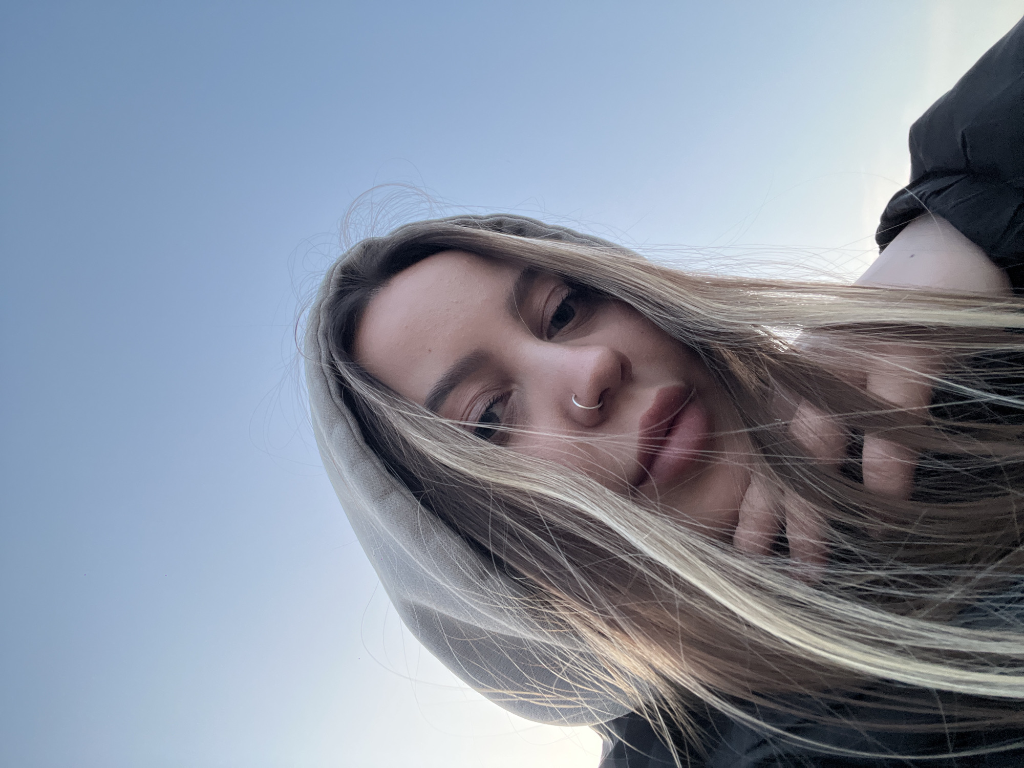 Captured in Novopokrovskaya, Russia, this low-angle image presents a close-up of a young woman's face and upper body against a clear, light blue sky. She has long, straight blonde and light brown hair with highlights, with some strands artfully blowing across her features. Her left eye is visible, and a silver hoop nose piercing adorns her left nostril. She wears a light-colored hooded sweatshirt, with the hood up, and a dark jacket. Her left hand is subtly visible near her mouth, partially obscured by her hair. Her expression appears calm or pensive, with a direct but soft gaze towards the camera. The outdoor setting and clear sky suggest daytime, possibly with a gentle breeze. There is no visible text.