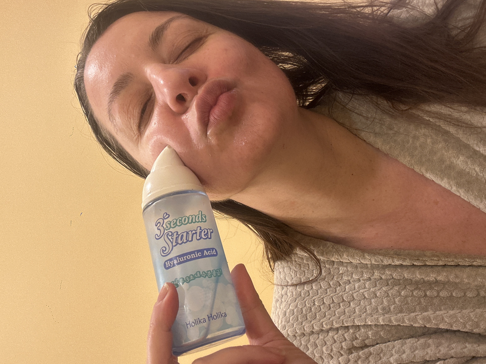 An eye-level shot captures a woman engaging in a skincare routine, seen from a low angle looking upwards. The primary subject is a woman with long, dark hair, whose face is prominent in the frame. Her eyes are closed, and her lips are pursed in a puckering or kissing gesture. Her skin appears dewy and well-moisturized.
She is holding a translucent light blue bottle with a white cap, identified as a skincare product, near her chin and mouth. The bottle's label clearly displays "3 seconds Starter" in light green and purple text, followed by "Hyaluronic Acid" and "Holika Holika" at the bottom, along with some Korean text.
The woman is wearing a light beige or cream-colored bathrobe with a textured, possibly waffle-knit pattern. The background is a plain, solid light yellow-beige wall or surface, providing a clean and uncluttered setting. The overall impression is one of self-care or product application, with a relaxed and possibly playful mood suggested by her facial expression. The scene is indoors, and the lighting is soft and even. While the context is Riga, Latvia, there are no specific visual cues within the image to indicate this location.