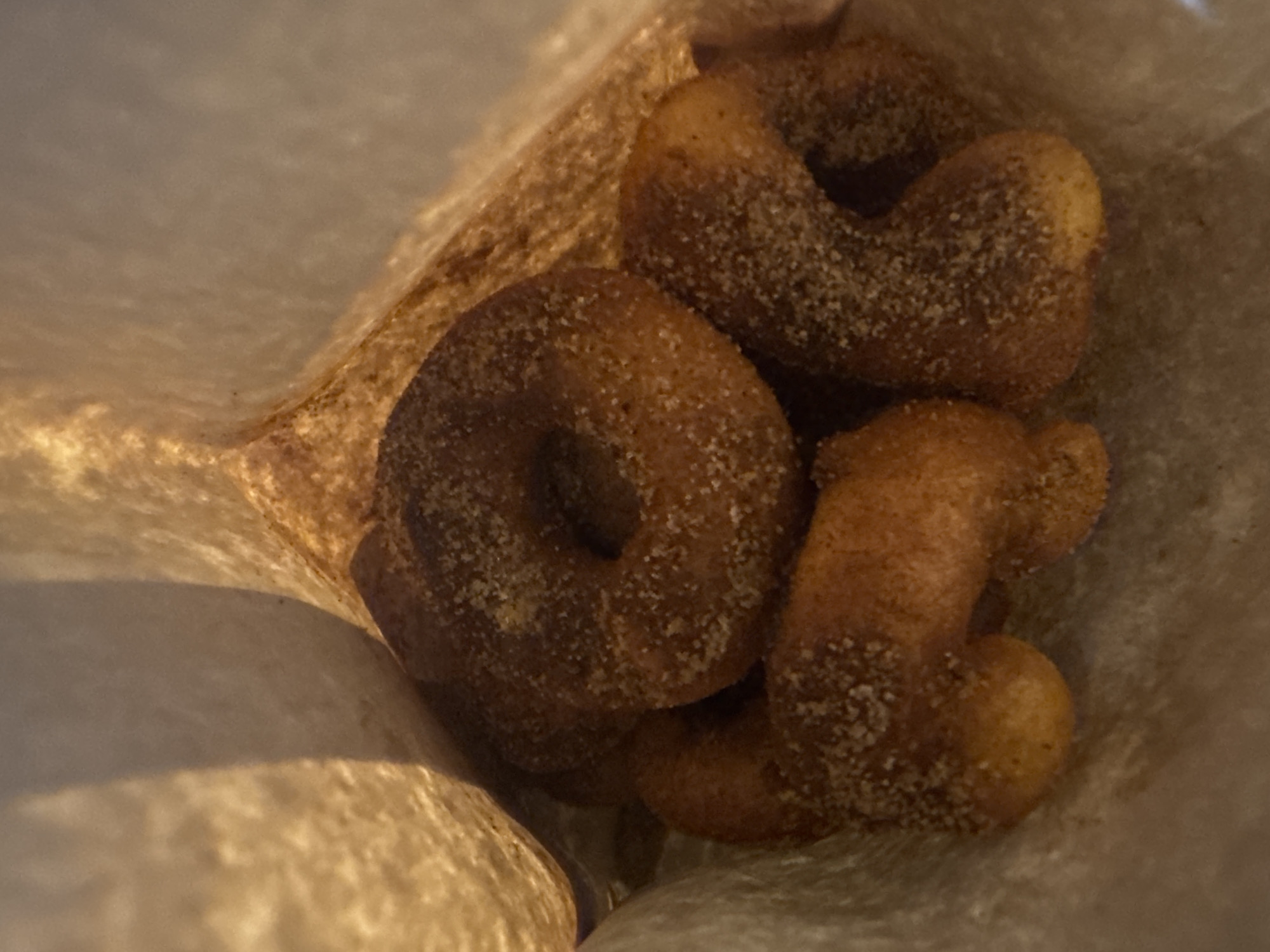 This close-up image captures several small, golden-brown pastries inside a light brown paper bag. The subjects are these individual pastries, which resemble mini doughnuts or churro bites. They are coated generously with a fine, granular mixture, likely sugar and cinnamon, giving them a speckled texture.
The objects visible are the pastries themselves, which vary in shape; some are distinctly ring-shaped, while others are irregular, elongated, or curved. These shapes suggest they might be doughnut holes or segments of fried dough. The container is a creased, plain paper bag, appearing slightly textured and potentially stained from the pastries.
The scene is focused tightly on the food item within the bag, looking down into it. The setting is implied to be a casual food context, perhaps a take-away or snack. No specific activity or event is taking place beyond the presentation of the food.
Notable details include the appetizing appearance of the sugary, fried snacks. The lighting appears warm, casting some subtle shadows and highlighting the sugar crystals on the pastries. There are no visible people, specific time of day cues, or weather details. No visible text is present on the pastries or the bag. Although not visually evident in the image, the provided context indicates this scene is located in Cape Town, South Africa.