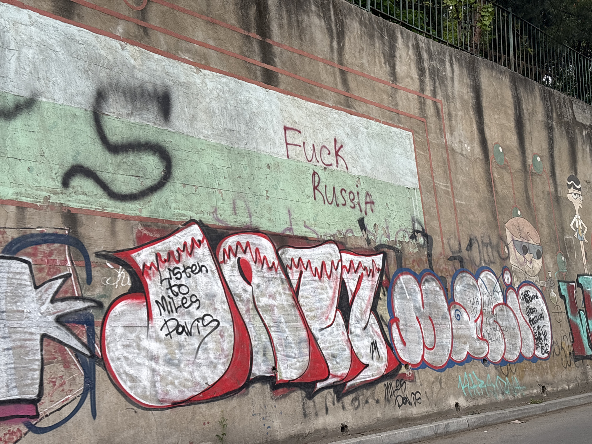 A concrete retaining wall, heavily marked with graffiti, is depicted in daylight. The wall features horizontal painted strips: a lower faded green band and an upper off-white band, both outlined in red. Above these strips, the concrete is bare and stained with dark vertical streaks. A black metal fence and green foliage are visible at the very top of the wall.
In the upper section, a large, stylized black spray-painted 'S' appears on the left. To its right, across the faded green and white bands, the words "Fuck Russia" are written in red spray paint.
The lower half of the wall is dominated by large, elaborate bubble-style graffiti letters, primarily in white with red and black outlines. These letters appear to spell "JAZZ MUSIQ".
Within the first large letter, "J", smaller black text reads "listen to Miles Davis". Below this main graffiti, also in black, are the words "Miles Davis".
Further right, within the main letters, smaller, less legible text is present, including repetitions of "TIVA VIVA" and phrases like "younG" and "from".
To the right of the main graffiti, several cartoon-like characters are drawn. One resembles Dexter from "Dexter's Laboratory," wearing sunglasses and holding a flask. Another figure, partially visible on the far right, appears to be Rick Sanchez from "Rick and Morty."
The wall shows signs of wear, peeling paint, and general urban decay. A concrete curb and a narrow strip of pavement are visible at the bottom of the image, suggesting a roadside or pathway setting. The lighting indicates it is daytime.