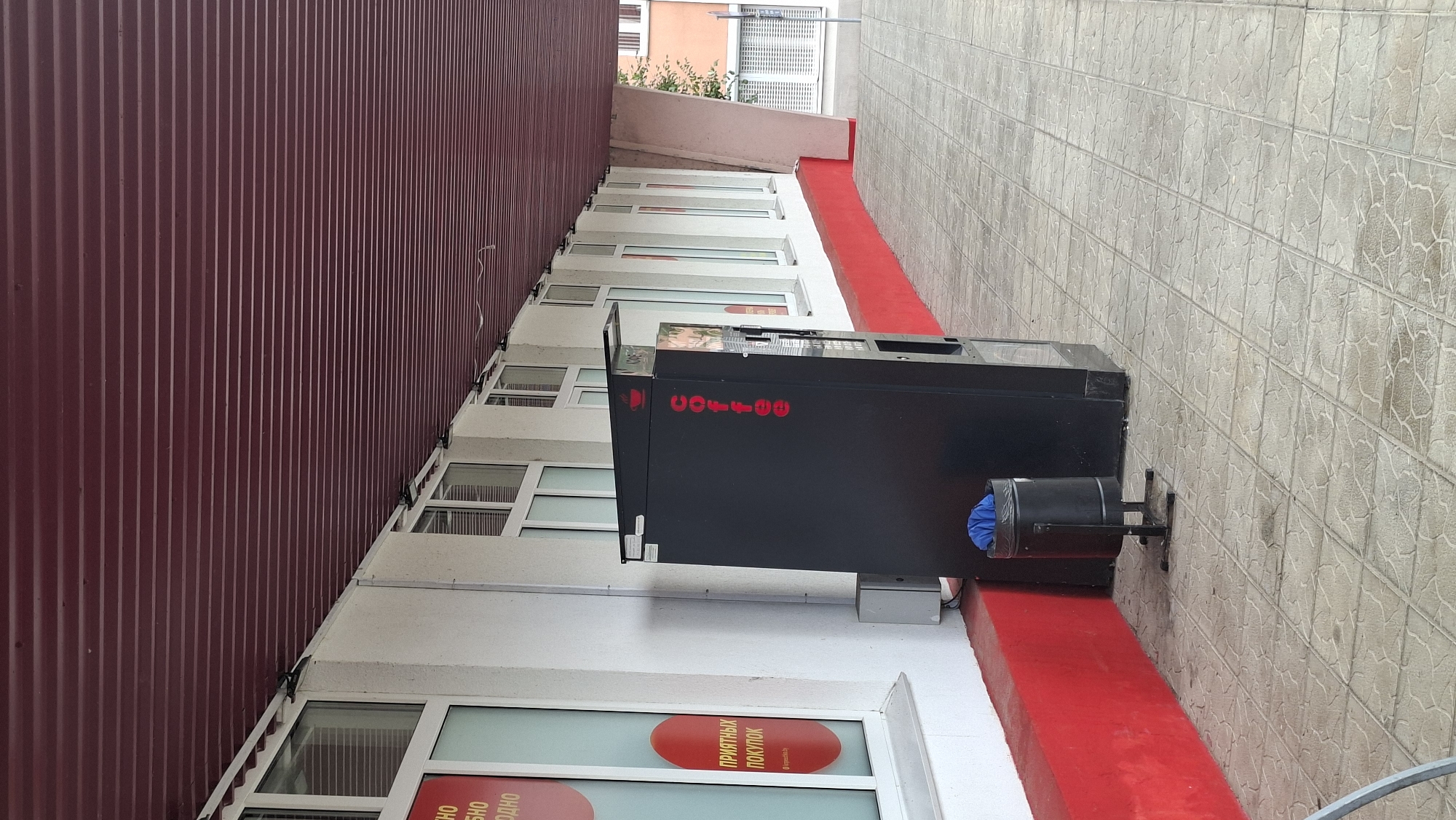 The image shows an outdoor scene in front of a building with a red metal roof extending over a walkway. The building has white walls and a red base. Along the wall are several large windows with white frames. To the left, a red circular sticker with white text is visible on one of the windows. In the center of the image, a tall black vending machine is positioned on a tiled walkway. The vending machine has red lettering that spells "coffee" vertically on its side, and a red coffee cup logo at the top. Next to the vending machine, to its left, is a black trash can with a blue bag inside. The walkway is paved with light-colored square tiles that appear wet. The background shows more of the building and some greenery further down the walkway.