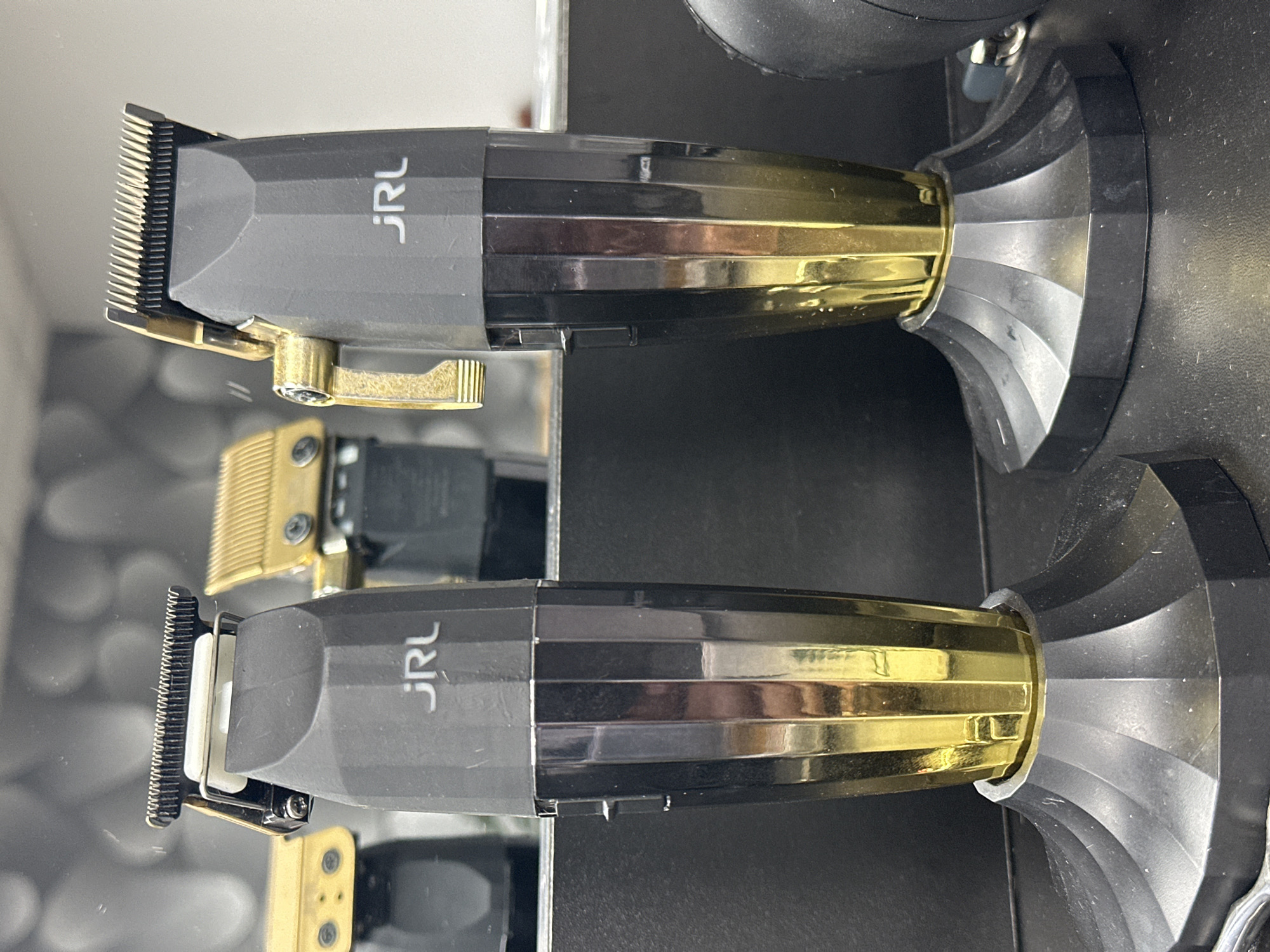 The image shows two professional hair clippers with black and gold accents, each resting on a black base. The clippers are positioned in front of a reflective surface, likely a mirror, which shows reflections of similar clippers and a blurred background. The clippers themselves have faceted bodies that transition from black to gold towards the base, and gold-colored blades and lever mechanisms. The brand "JRL" is visible on the body of each clipper. The setting appears to be a barbershop or salon. There is no indication of the specific location within Uzhhorod, Ukraine, as the image only contains objects and their immediate surroundings.