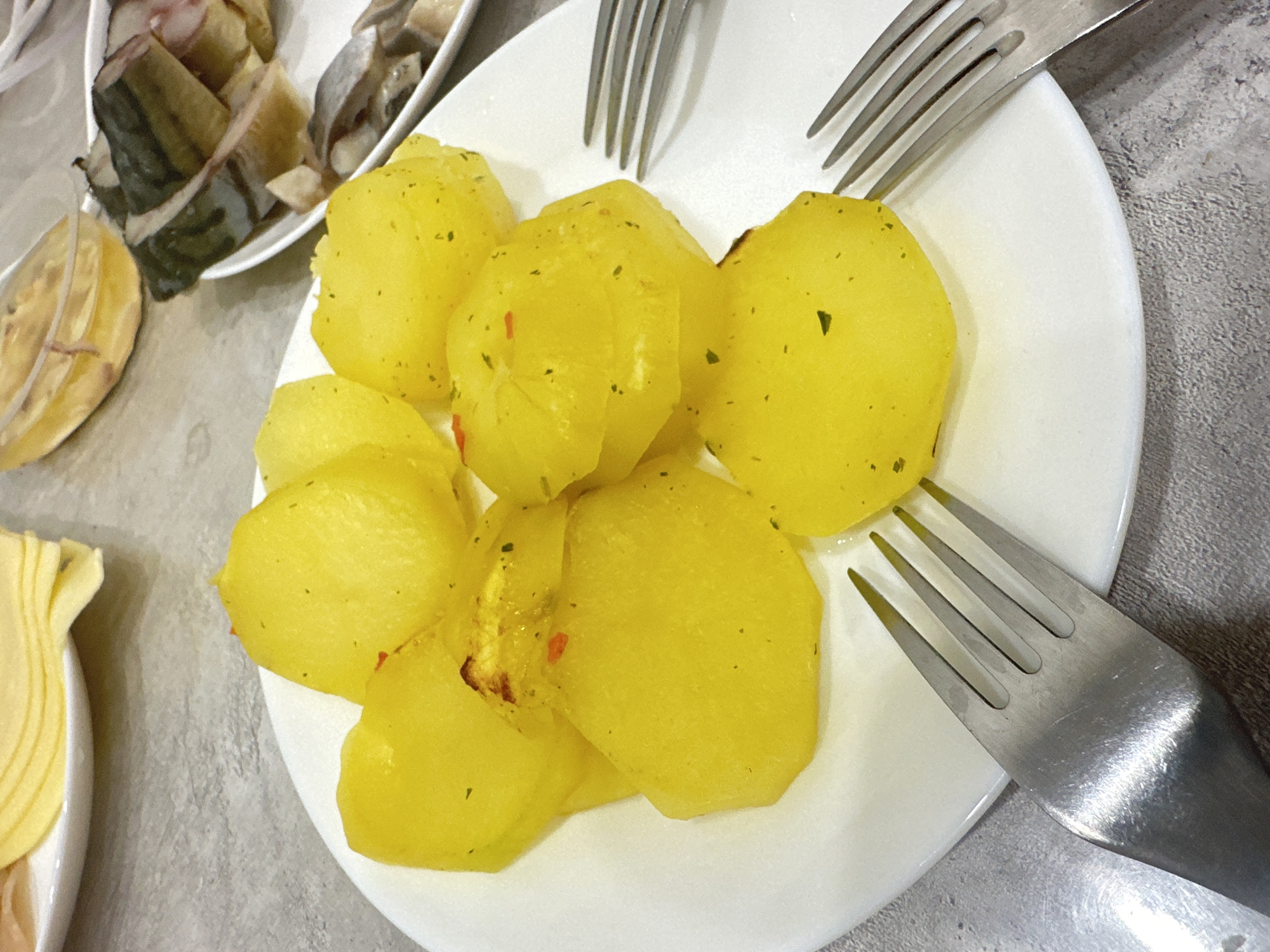 A close-up shot depicts a white plate containing several pieces of boiled potatoes, seasoned with herbs and spices. Three forks are laid across the plate, their tines pointing towards the potatoes. To the left, a separate plate holds pieces of fish, and a side dish that appears to be sliced cheese. The setting suggests a dining environment. The precise location within Chaikovsky, Russia, is not discernible from the image alone.