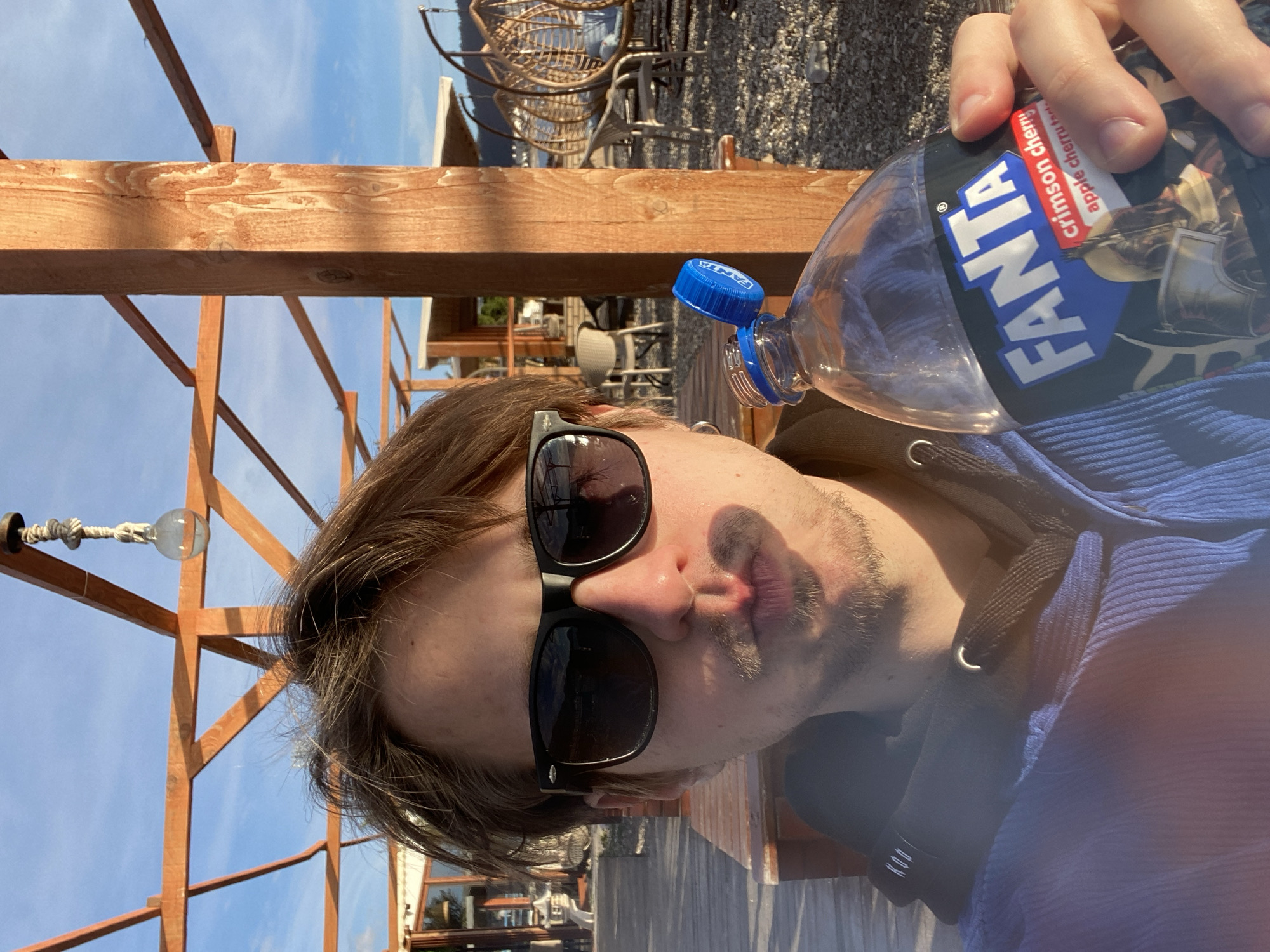 A selfie-style image shows a person holding a nearly empty bottle of Fanta Crimson Cherry soda outdoors. The person has brown hair and is wearing sunglasses and a blue hoodie. The background reveals a wooden structure with hanging lights against a blue sky, suggesting an outdoor patio or deck. In the distance, there are wicker chairs, gravel, and glimpses of buildings. The setting appears to be a sunny day in Bar, Montenegro, as the backdrop includes elements characteristic of a coastal town like Bar.