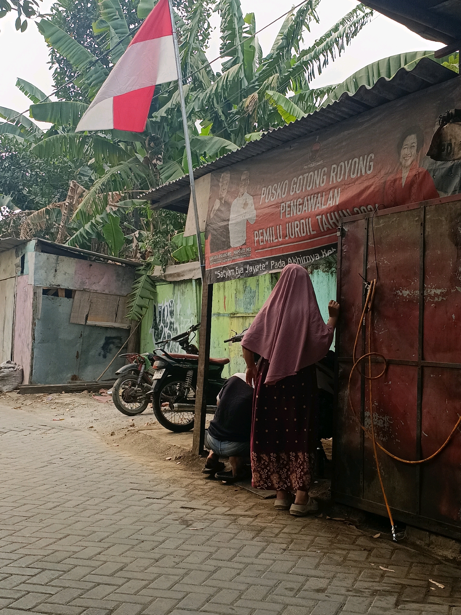 The image captures an outdoor scene in Tangerang, Indonesia, likely a street in a more rural or less developed area, given the corrugated metal roofs and simple building structures. A red and white Indonesian flag is prominently displayed on a metal pole in the upper left portion of the frame, suggesting a patriotic or official context.
In the foreground, a person wearing a pink headscarf and a patterned maroon dress stands with their back to the camera, facing a large, weathered red metal door. Their right hand is extended, appearing to be in the process of knocking or interacting with the door. A coiled yellow hose is attached to the doorframe, hanging downwards.
To the left of the person, partially obscured, is a seated individual wearing dark clothing and light-colored pants. A dark-colored motorcycle with a brown seat is parked behind them. Further to the left, a small, weathered building with faded pastel-colored walls is visible. The ground is paved with interlocking bricks, and scattered debris suggests a somewhat untidy environment.
A large banner is stretched across the top of some structures behind the flag. The banner is predominantly red and features text in white and black, including "POSKO GOTONG ROYONG PENGAWALAN PEMILU JURDIL TAHUN 2024," which translates to "Mutual Cooperation Post for Fair Election Monitoring 2024." Images of two individuals are also present on the banner. Banana trees and other foliage are visible in the background, contributing to a tropical atmosphere. The overall lighting suggests it is daytime, possibly overcast.