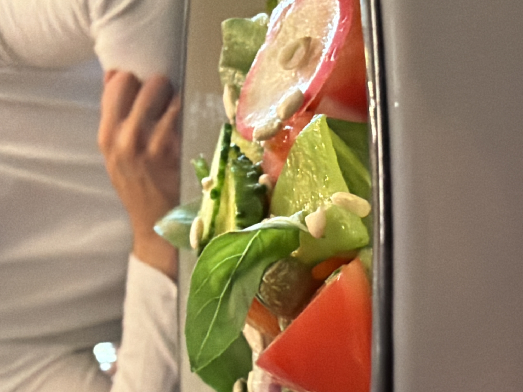 A close-up, low-angle photograph captures a salad in a bowl with a person in the background, set in Kazan, Russia. The foreground prominently features a vibrant salad contained within a light gray bowl with a dark rim. Visible salad components include a large, fresh green basil leaf centrally placed, alongside slices of red tomato, segments of green cucumber, and light green bell pepper or similar vegetable pieces. Scattered over the salad are numerous small, elongated, light-colored seeds, consistent with pine nuts or sunflower seeds. A small, round, brownish-green object, possibly an olive or caper, is also discernible. In the blurred midground, a person wearing a long-sleeved white garment is visible from the chest upwards, with their hands clasped or resting on a surface, indicating an seated posture at a table. The background is indistinctly blurred, showing general indoor lighting.