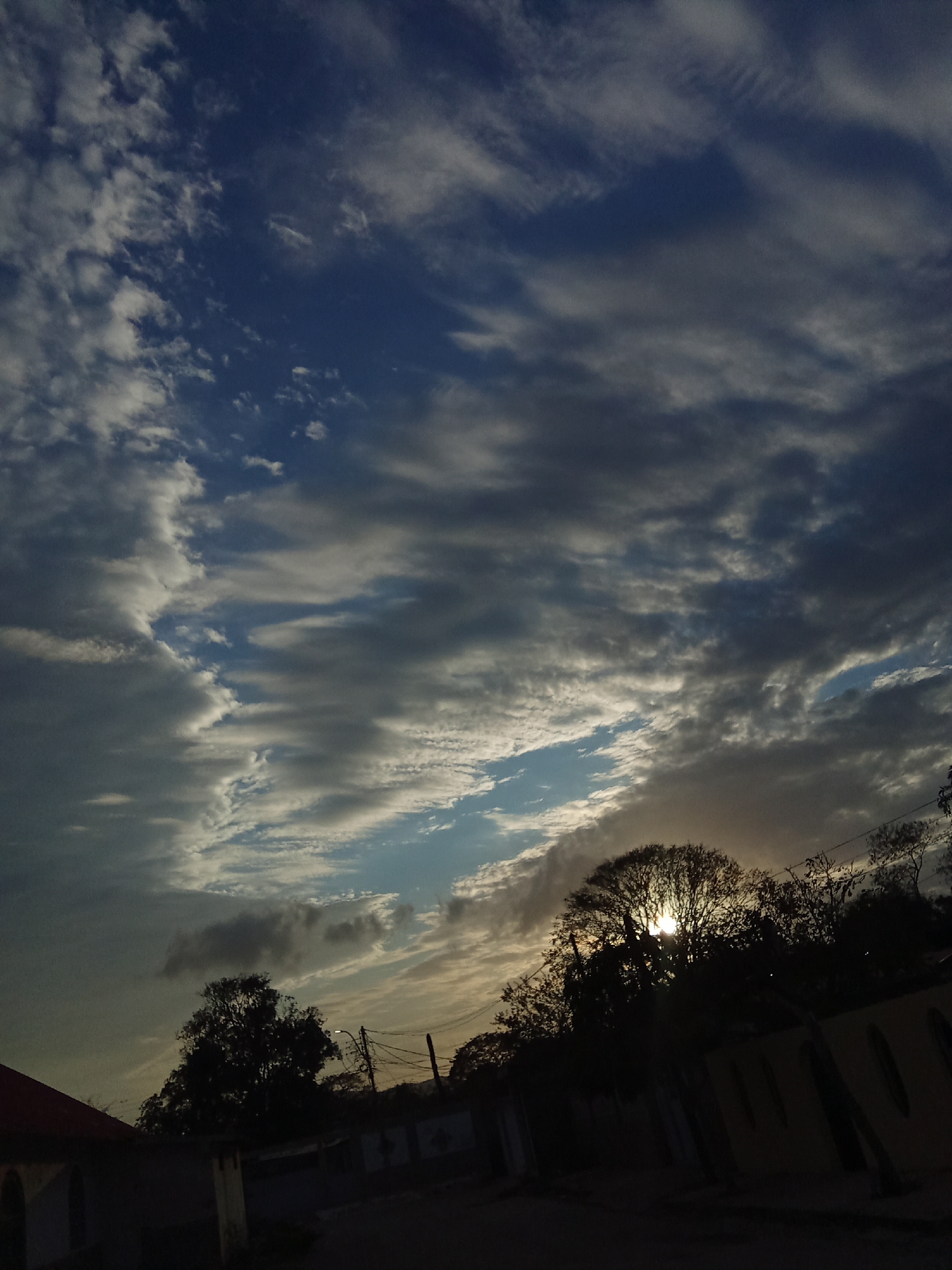 The image presents a sky filled with striated clouds, predominantly gray with streaks of blue peeking through. The horizon is defined by silhouetted trees and building structures, with the sun partially obscured by the foliage, creating a bright halo effect. In the foreground, there are building rooftops, walls, and what appears to be electrical wiring against the sky. Specific identification within Juan Griego, Venezuela, is not discernible from the provided visual data.
