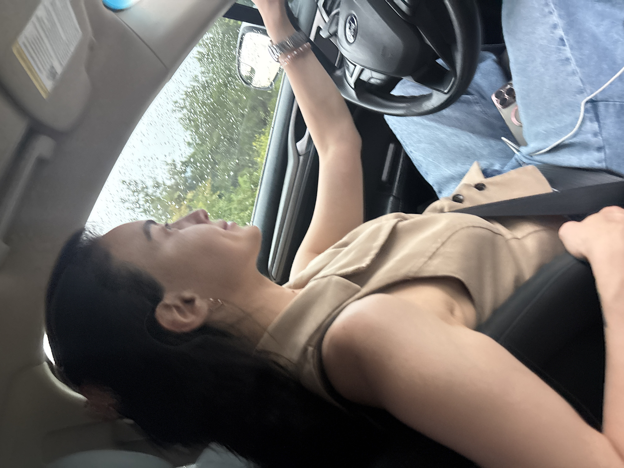 The image shows a woman driving a car in what appears to be a city. The woman has dark hair and is wearing a beige sleeveless top and blue jeans. She is looking forward, and her hands are on the steering wheel. The car's interior is visible, including the dashboard and a smartphone. The exterior of the car shows a blurry view of trees and what appears to be raindrops on the windshield, suggesting it might be raining. The scene does not contain any specific landmarks or identifying features that would allow for pinpointing the location to Tbilisi, Georgia.