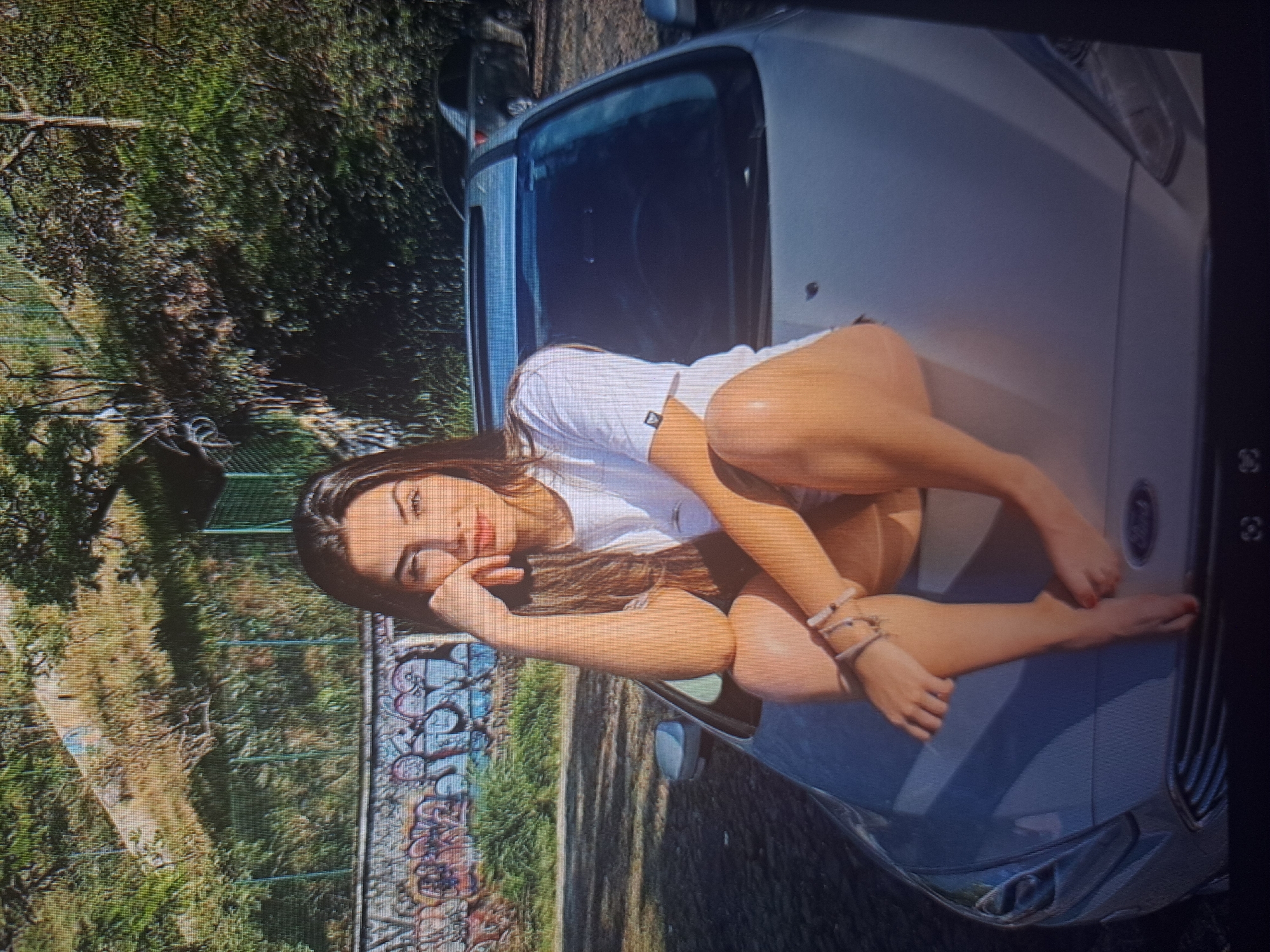 A young woman with long dark hair, wearing a white t-shirt, is sitting on the hood of a silver Fiat car. She is resting her head on her hand and looking at the camera. The car is parked on a street next to a fence covered in graffiti. Behind the fence is a grassy area with trees. The location appears to be an urban environment, potentially a park or roadside in Yerevan, Armenia. The scene suggests a casual setting, possibly a spontaneous photo opportunity.