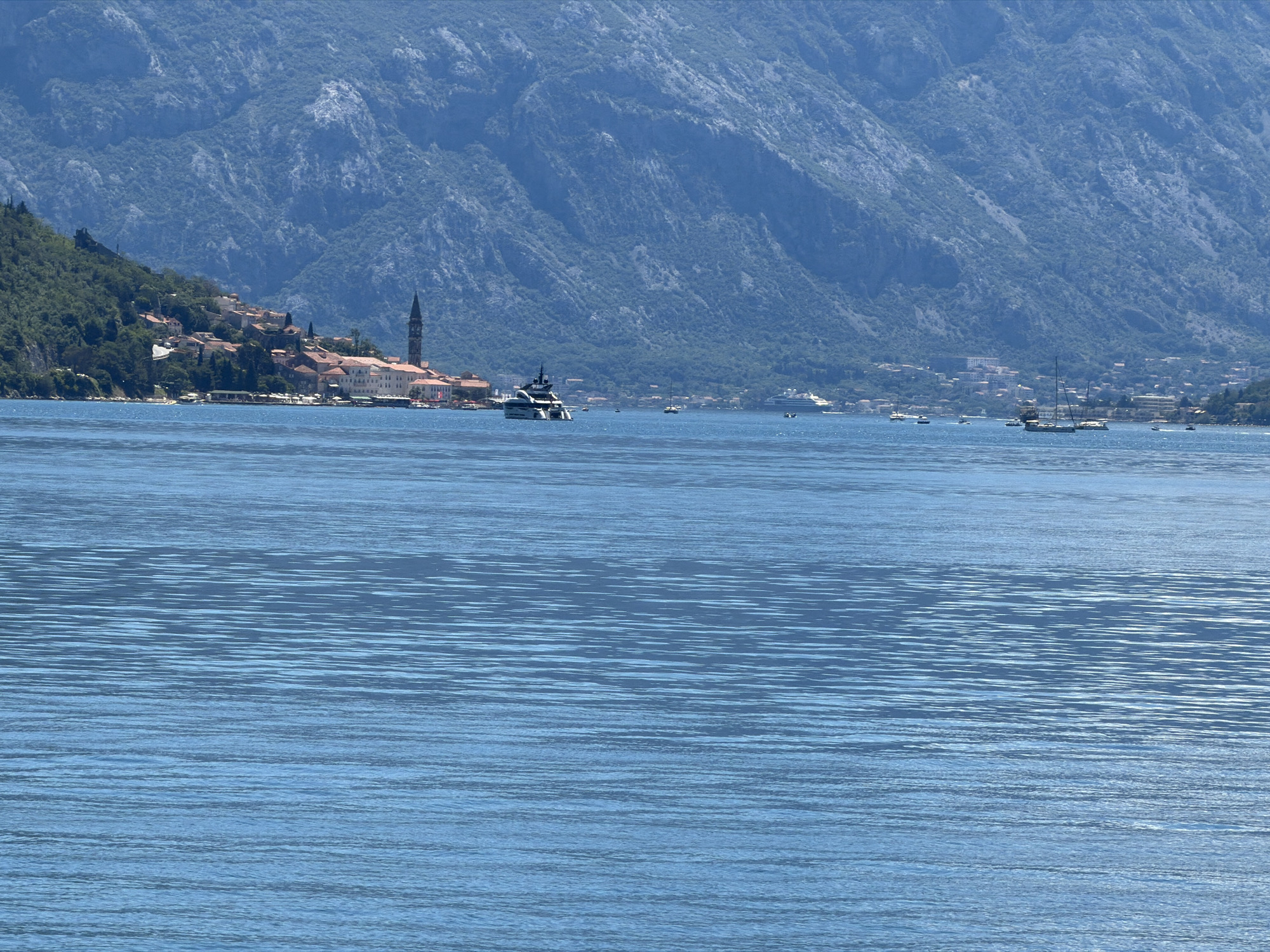 This image captures a wide-angle view of the Bay of Kotor in Montenegro, likely near the town of Lipci, as indicated by the distinctive mountainous backdrop and coastal settlement. The foreground is dominated by a calm expanse of blue water, showing gentle ripples that reflect the bright sunlight.
In the mid-ground, a picturesque coastal town is nestled at the foot of a steep, forested mountain. The town features a cluster of red-tiled roofs and light-colored buildings, with a prominent church steeple rising above the rooftops. Several boats are visible on the water, including a large, sleek yacht with multiple levels, a sailing ship with tall masts, and smaller motorboats. Further out, a larger white vessel, possibly a cruise ship or ferry, can be seen.
The background is dominated by a massive, rugged mountain range, its slopes covered in dense green vegetation. The texture of the rock faces and the scattered trees are discernible, emphasizing the imposing scale of the natural landscape. The overall scene conveys a sense of tranquility and the beauty of a coastal European setting. There are no discernible people in the image.