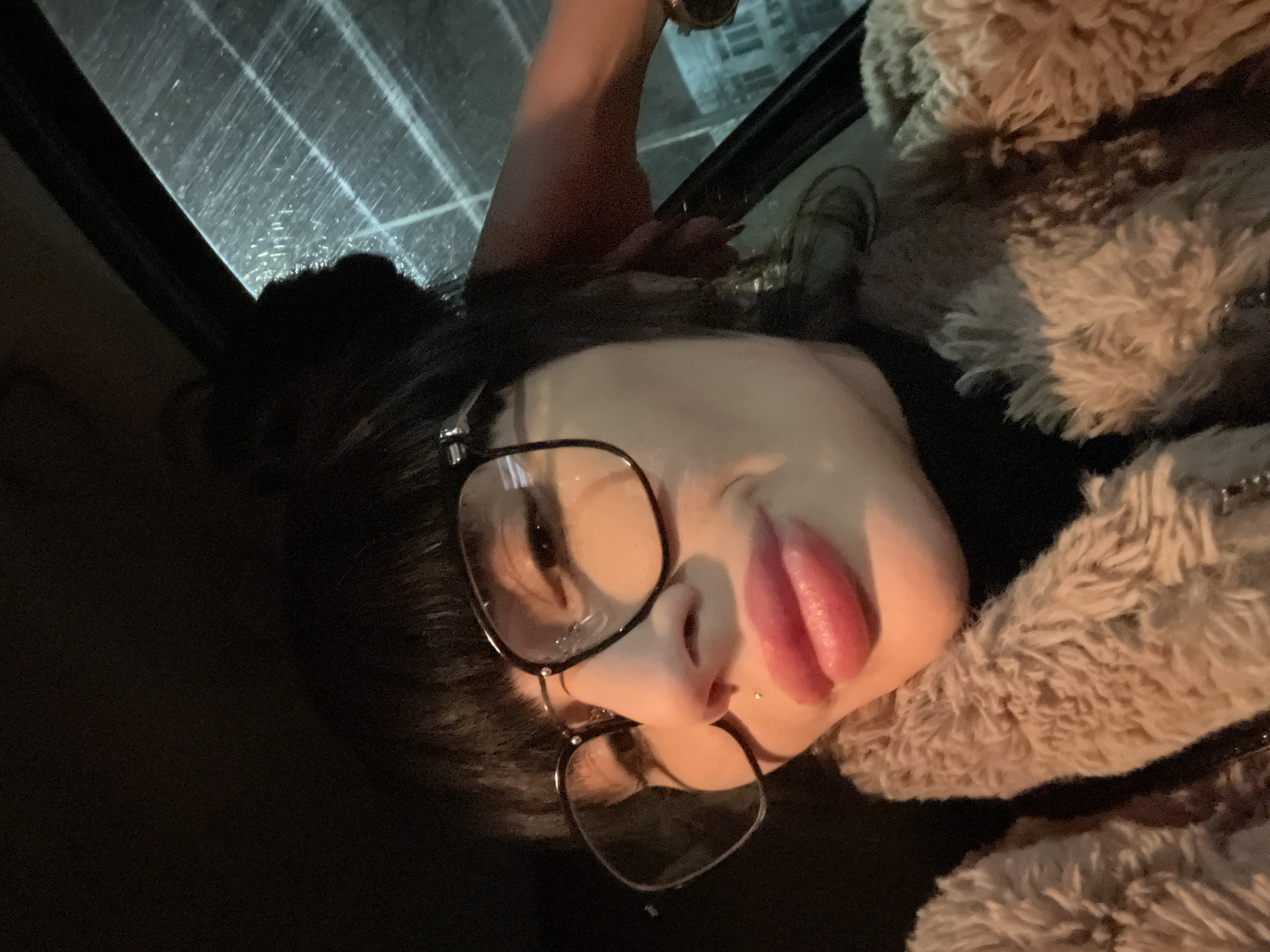 The image is a close-up selfie of a person with dark hair wearing black glasses and a furry coat. The person's face is tilted, and their lips are pursed. They appear to have a lip piercing. The background suggests they are indoors, possibly in a car, as a rain-streaked window frame is visible. The lighting is dim. The image does not provide any information that indicates a location in Varna, Bulgaria, or any other specific location.