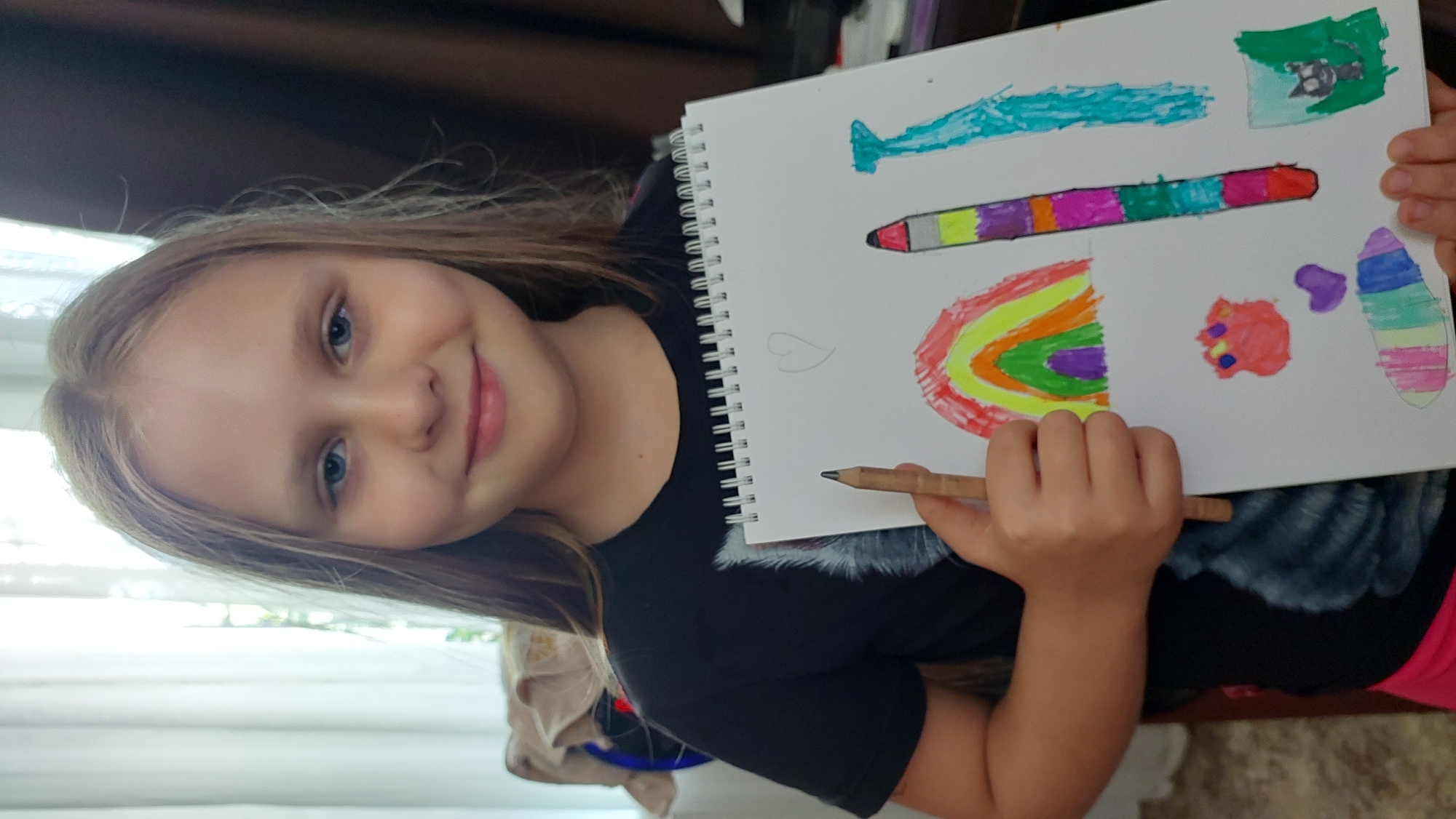 A young girl with blonde hair and blue eyes is holding up a drawing in a spiral-bound notebook. She is wearing a black t-shirt with a wolf graphic. The drawing contains a rainbow, a tall crayon-like object with colorful stripes, and a small black cat in a green landscape. She is smiling at the camera. The background shows a curtain and a blurred chair. This image does not provide any information about the location in Lermontov, Russia.