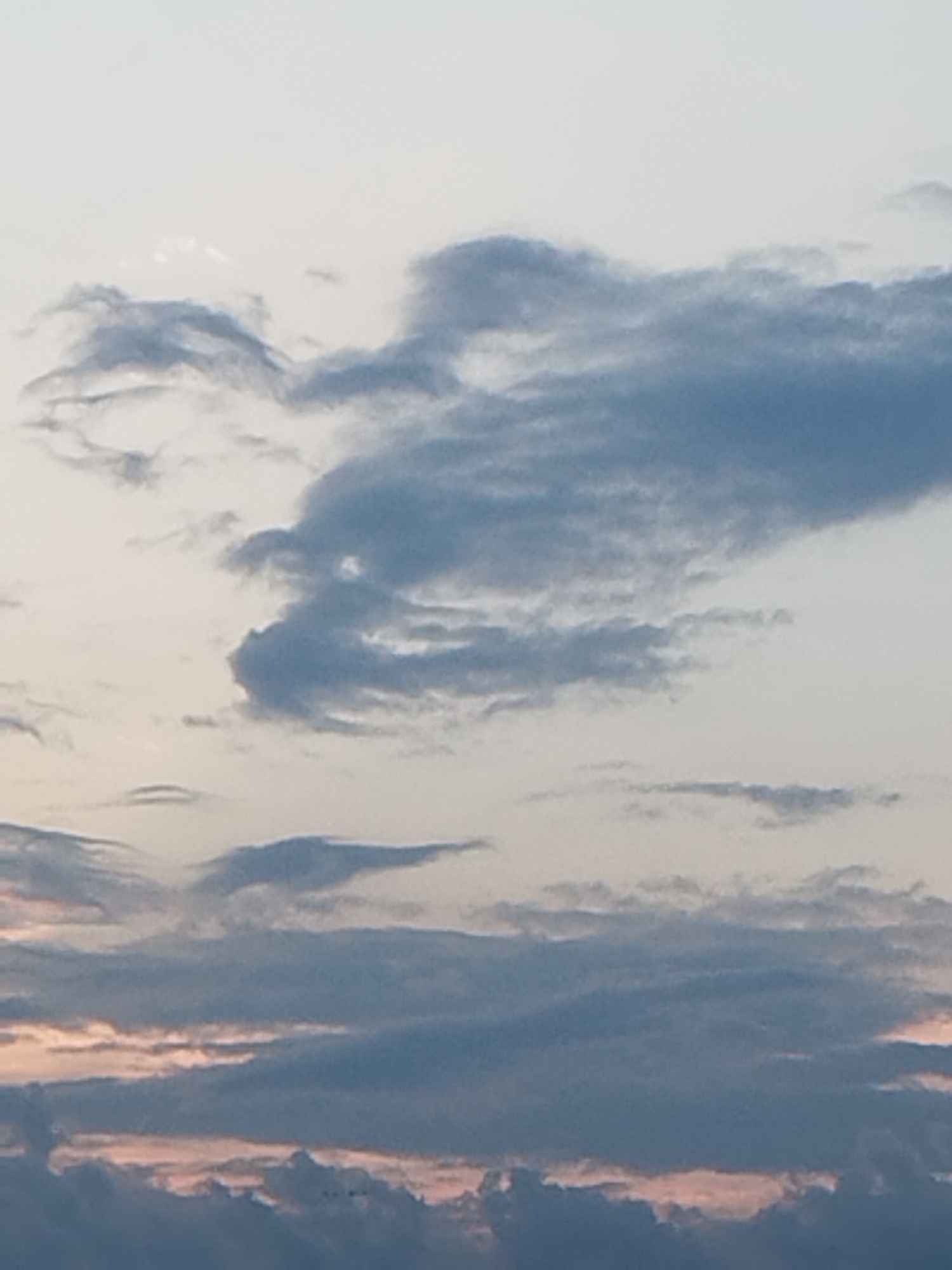 The image is a photograph of clouds in the sky. The clouds are varied in shape and color, with some appearing dark and ominous, while others are lighter and more wispy. The sky itself is a pale blue, with hints of pink and orange near the horizon, suggesting it may be dawn or dusk. There are no visible people, subjects, or objects in the image, nor any actions or interactions. The environment is purely atmospheric. The location in Bucharest, Romania, cannot be determined from the visual information provided.