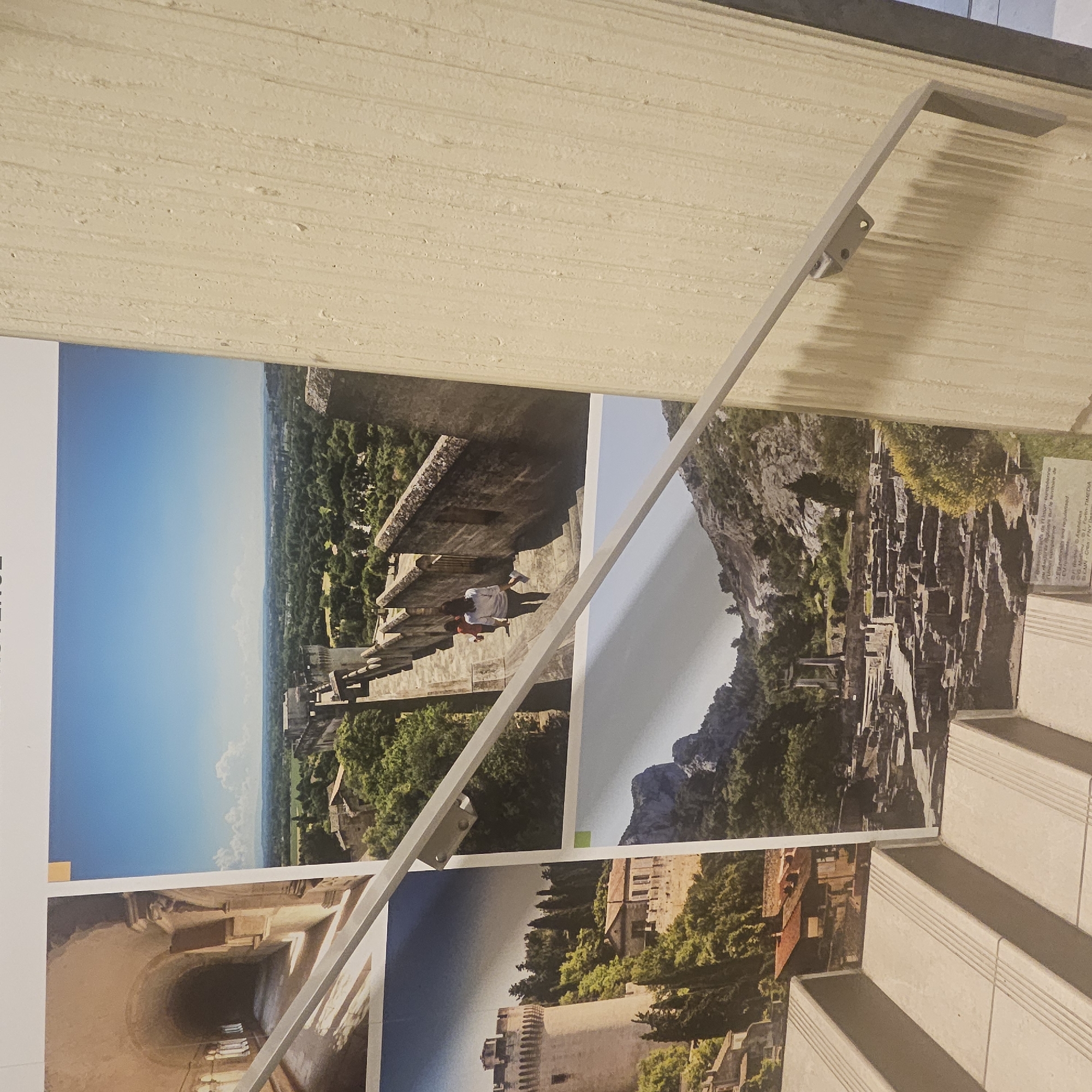 The image displays a collection of photographs, possibly part of an exhibition or informational display. The photographs are arranged in a grid-like fashion, with some overlapping. One photograph captures a view from atop ancient stone fortifications, overlooking a verdant landscape under a clear blue sky. Two individuals are seen walking down a stone pathway along the fortifications. Another photograph features a cylindrical stone tower and a cluster of buildings nestled within a hilly, tree-covered area. A third image depicts ruins of stone structures in a rocky, mountainous environment, with vegetation growing around them. The photographs are mounted on a white background. In the foreground, a light-colored staircase with multiple steps is visible, leading up towards the display. A metal handrail runs diagonally across the frame, attached to a textured, cream-colored wall. The text "PROVENCE" is visible at the top of the display, suggesting a regional theme. The location is identified as Lançon-Provence, France.