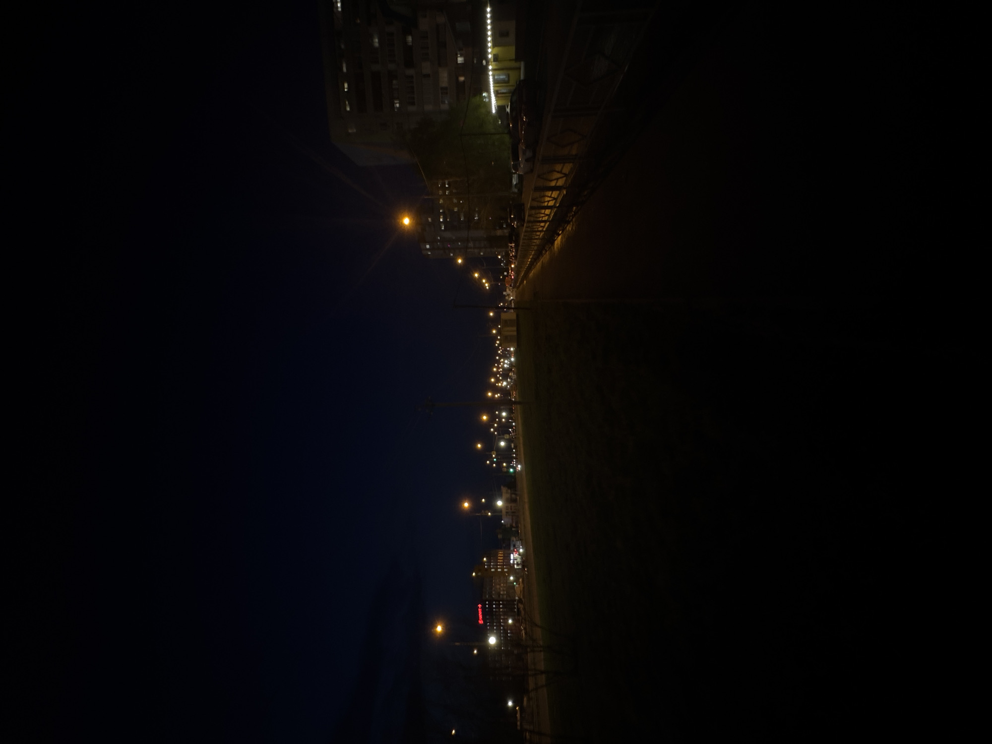 The image presents a nighttime scene of an urban street or boulevard, captured from a slightly elevated vantage point. The sky is dark, indicating late evening or night. A prominent feature is a long stretch of road illuminated by numerous streetlights, casting a warm yellow-orange glow that extends into the distance, creating a strong sense of perspective. Some lens flare is visible around the brightest light sources.
Multi-story buildings, likely residential or commercial, line the street, particularly on the left side of the frame (when viewed correctly). Many windows within these buildings are illuminated, suggesting activity. Interspersed among the buildings are dark silhouettes of trees or vegetation. On the right side, a strip of grass or a park area runs parallel to the street.
While no individual people are visible, the presence of vehicle lights in the far distance implies ongoing traffic and general city movement. The scene captures the typical ambiance of an active city at night. A notable detail is a red illuminated sign on a building in the mid-ground, clearly displaying the word "БИЛЛА" (BILLA), identifying it as a supermarket, and providing a specific location cue for an urban environment. The weather appears clear, with no precipitation or fog.
