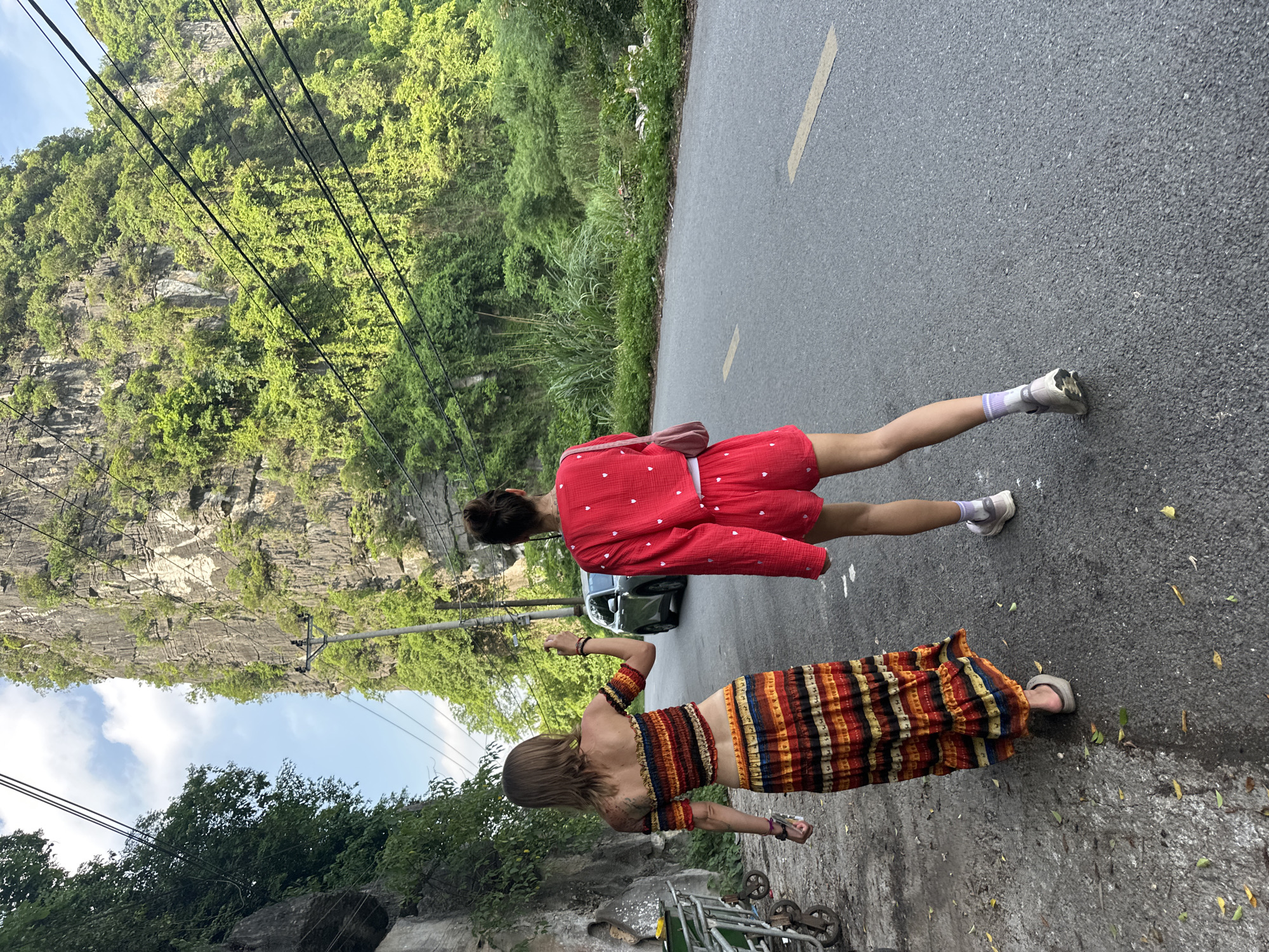 Two individuals are captured from behind, walking on an asphalt road. The person on the left wears a vibrant, striped, off-the-shoulder crop top and a matching flowing skirt, complemented by sandals. The individual on the right is dressed in a red outfit consisting of a long-sleeved top and shorts, paired with sneakers and socks. A light pink bag is slung over their shoulder. A dark-colored vehicle is visible on the road behind them. The environment features a steep, lushly vegetated rock face on the left, with a line of power lines running across the frame, and dense greenery on the right side of the road. The scene appears to be a scenic route in Nam Hoa Lư Ward, Vietnam, likely a mountainous or karst region, suggested by the imposing rock formation.