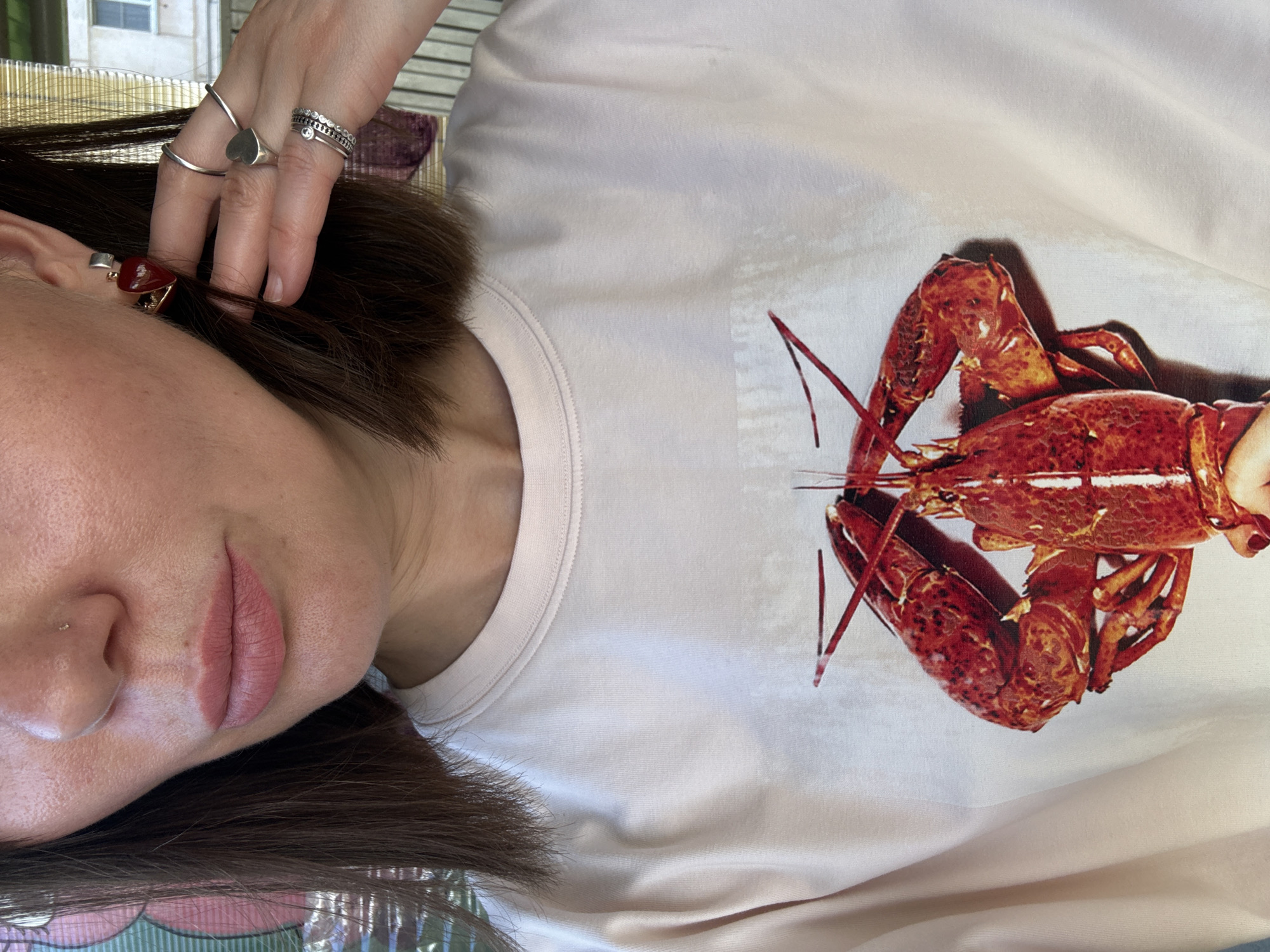 The image is a close-up shot of a woman wearing a light pink t-shirt with a graphic of two lobsters. The woman has fair skin, brown hair, and is wearing makeup. She has a nose stud and is wearing heart-shaped earrings and several silver rings on her right hand. The background appears to be an outdoor setting with a building and blinds visible. This image is not located in the city of Chaikovsky, Russia.
