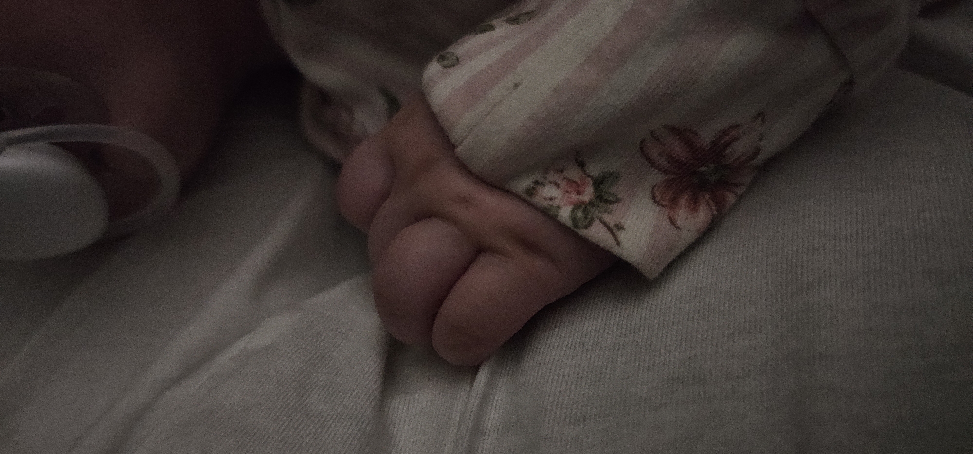 A close-up shot depicts a baby's hand holding onto a light-colored fabric. The hand is small, with visible skin folds and creases. The baby is wearing a pink and white striped sleeve with floral patterns. A pacifier is visible near the baby's face. The fabric appears to be a soft material, possibly a blanket or clothing. The setting is indoors, with subdued lighting.