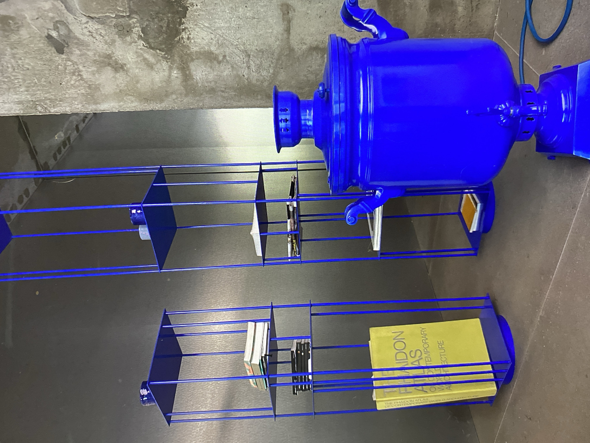 The image shows a bright blue samovar on a pedestal, placed in front of a textured concrete wall. To the left of the samovar are two tall, slender blue metal shelving units. The shelving units are open and contain books and what appear to be small decorative objects on their shelves. The larger shelving unit on the left has a yellow book with "PHaidon ATLAS" and "CONTEMPORARY WORLD ARCHITECTURE" visible on its spine, as well as stacks of other books. The overall scene is indoors, with a dark grey wall in the background of the shelving units. The location is identified as Khimki, Russia. There are no people visible in the image.