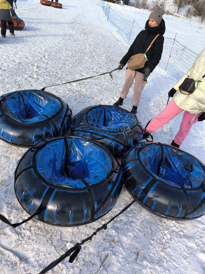 The scene depicts a snow-covered slope at a recreational facility in Prybuzke, Ukraine, under clear daylight conditions. Four black and blue snow tubing inner tubes are arranged in the foreground on the snow. Each tube has a visible blue interior and black exterior with black webbing and handles. Two female individuals are present, standing near the tubes. One woman, positioned centrally, wears a gray knitted beanie, a black puffer jacket, pink pants, black boots, and gray gloves; she has a tan cross-body bag and holds a long green strap connected to the lead tube. The second person, partially visible on the right, wears a light-colored puffer jacket, pink pants, and black boots; they have a dark cross-body bag and their right foot is near one of the tubes, with a black strap extending from it. Both individuals appear to be preparing or pulling the connected snow tubes uphill. In the background, a blue safety net runs along the right side of the slope, and other individuals on orange snow tubes are visible further up the incline.