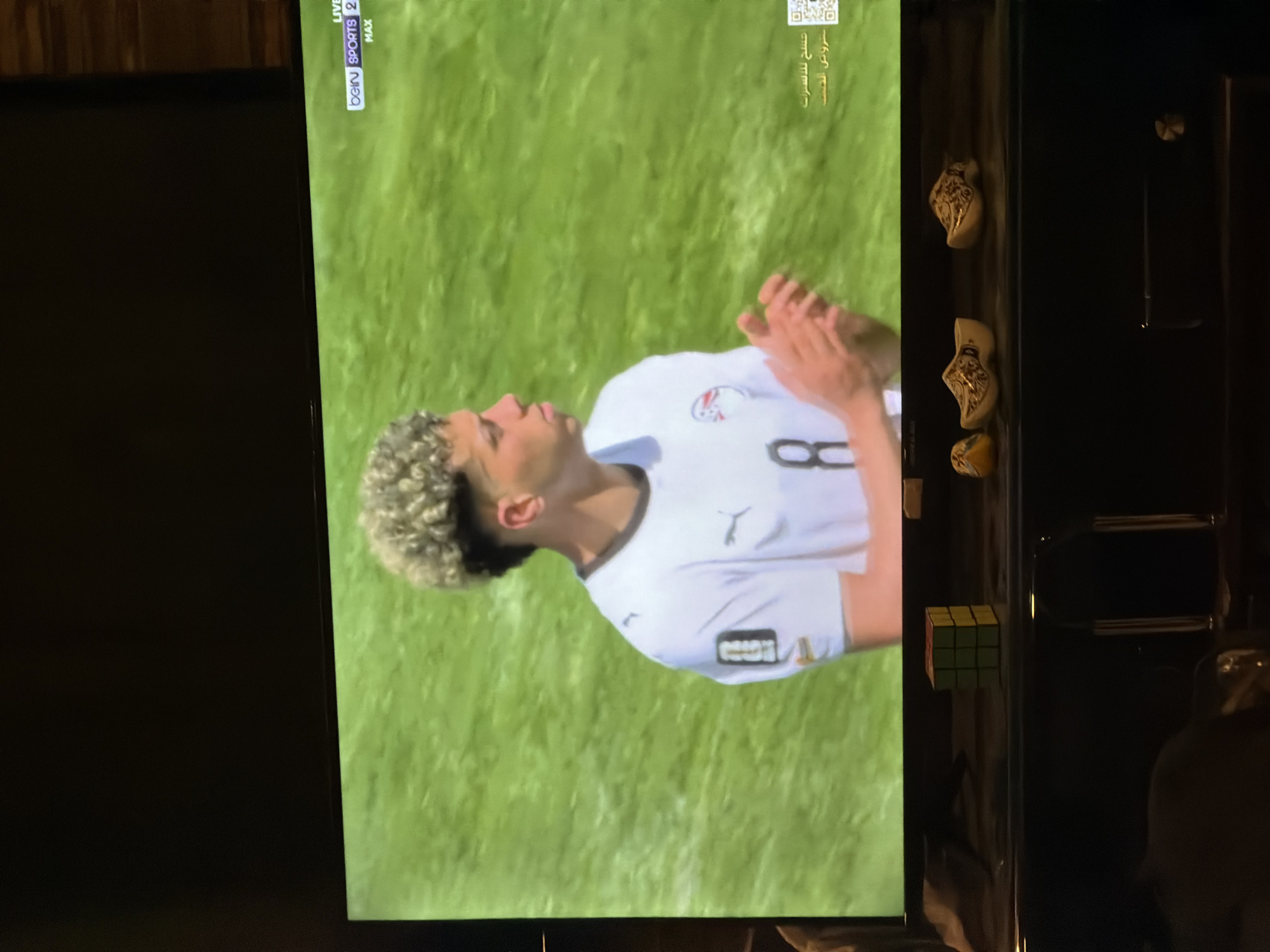 The image shows a television screen displaying a soccer match. A young male athlete in a white jersey with the number 8 is on the field. He has blonde, curly hair and is looking to his right. The background is a green field. On the bottom shelf of a furniture unit below the TV, there are three decorative clogs, a Rubik's Cube, and other objects that are not clearly identifiable. The furniture unit is dark brown. The overall scene appears to be a living room or entertainment area. The location of Sarouja Municipality, Syria, is not visually evident in the image.
