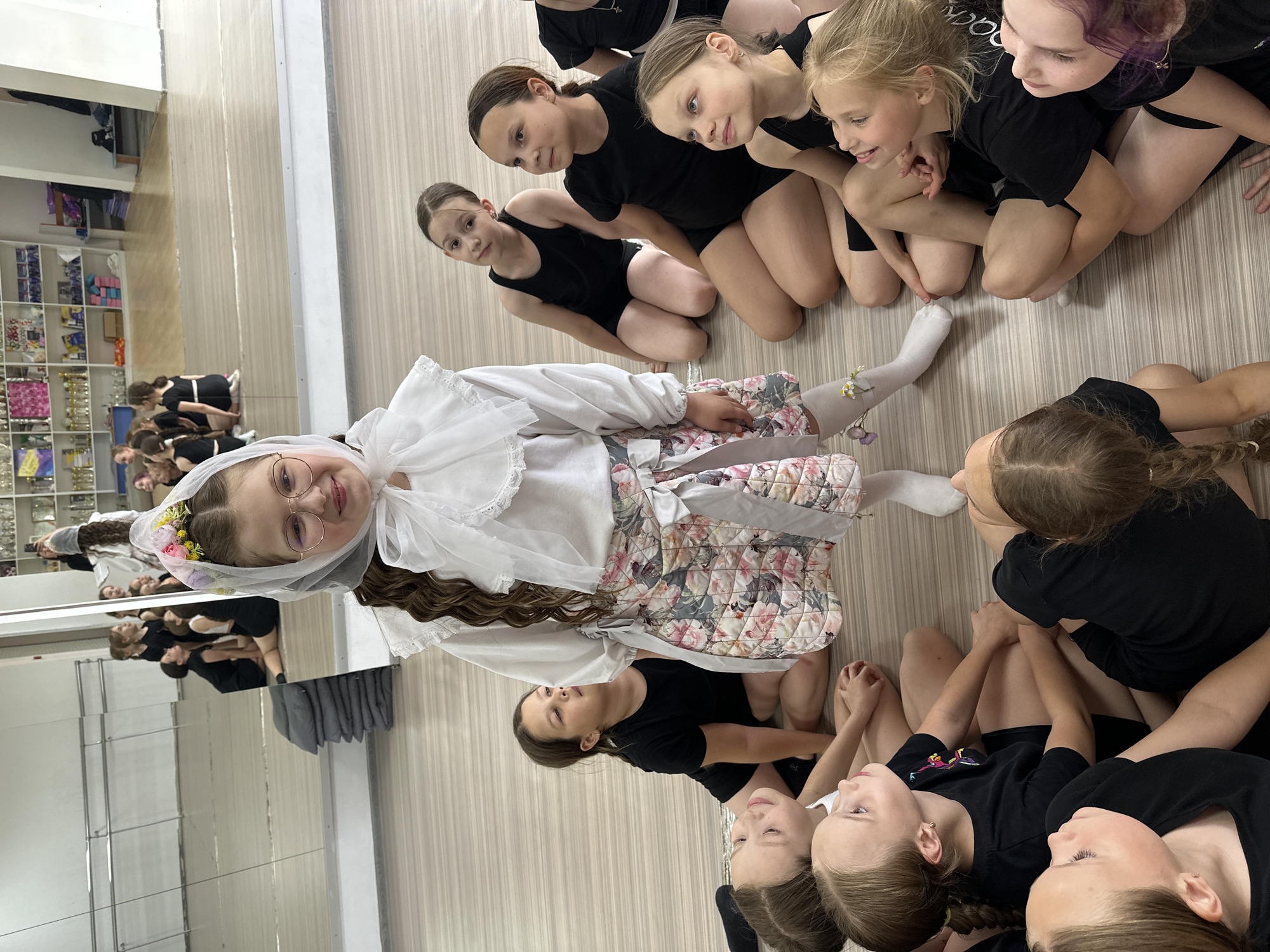 A wide-angle indoor shot depicts a group of young girls in a dance studio, likely in Votkinsk, Russia. The image is oriented with the horizontal axis slightly tilted. A young girl in a light-colored costume with a floral headpiece and glasses is lying on the floor, facing the camera. She has light brown hair and is smiling. She is surrounded by a group of girls in black leotards, who are sitting on the floor and looking at her. The floor is a light brown color, possibly linoleum or wood laminate.
There is a large mirror on the wall behind the group, reflecting the scene. In the reflection, more dancers in leotards can be seen. The room appears to be a dance studio with dance equipment and storage shelves visible in the reflection. The lighting is bright and even.