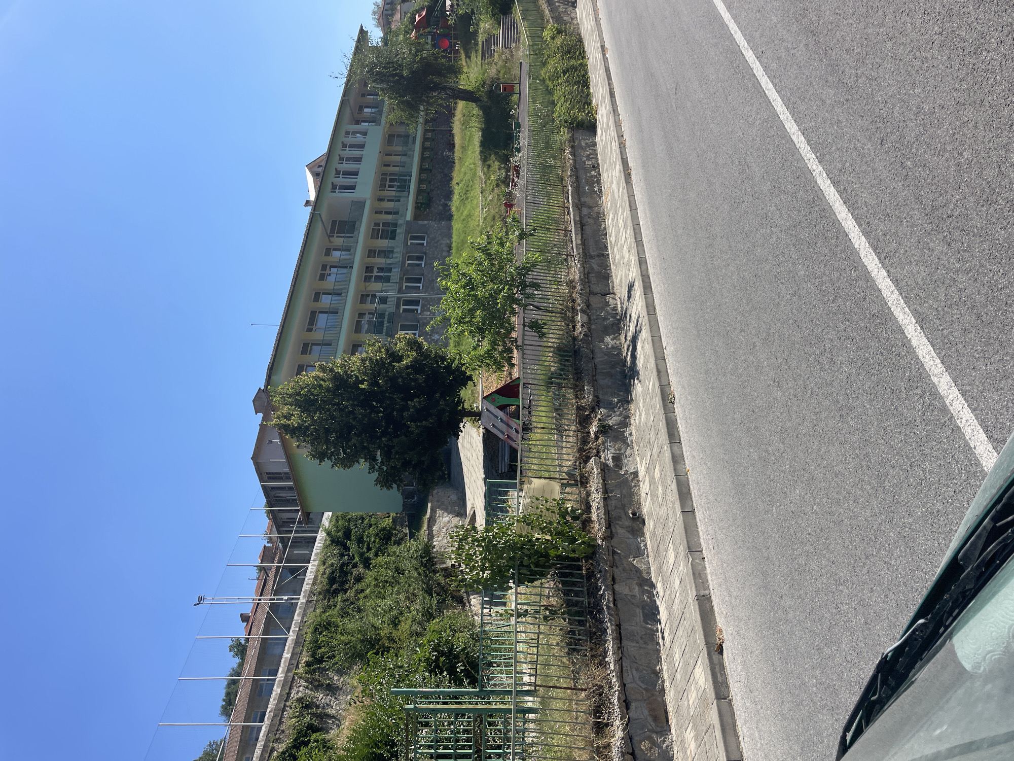 The image depicts a modern, light green, three-story building with numerous windows and balconies, possibly a school or dormitory, situated on a hillside. In front of the building, there is a fenced-off playground area with a small red and green play structure. A paved walkway runs alongside the building and playground, bordered by a stone retaining wall. A paved road with a white line marking is in the foreground. Lush green trees and foliage surround the building and playground. The sky is clear and blue. The image was taken in the city of Oryahovets, Bulgaria.