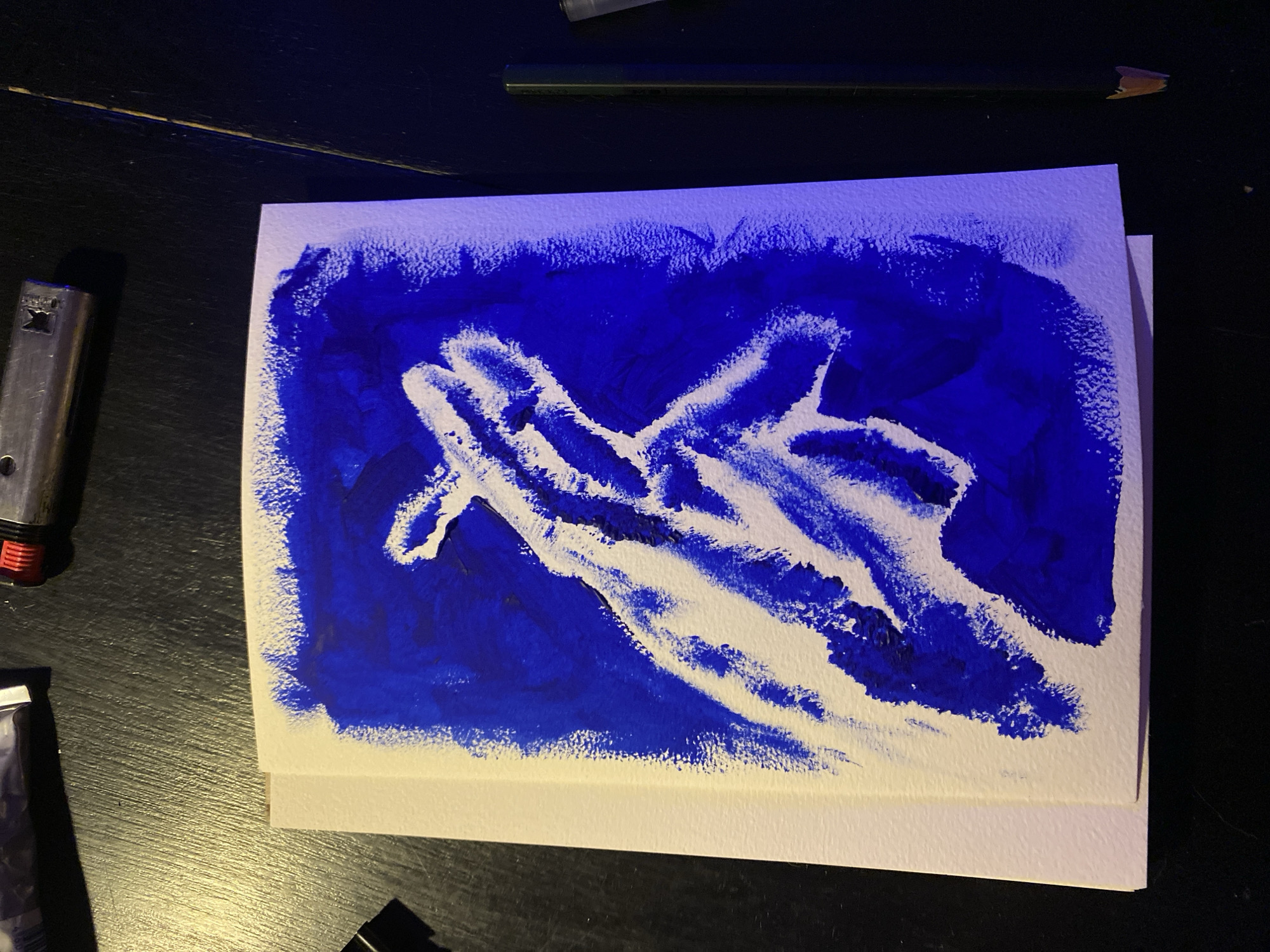 An overhead shot shows a vibrant blue and white abstract painting resting on a dark, textured table in Moscow, Russia. The artwork, on white paper, features a textured, rich blue background, against which a large, irregular white shape resembling an outstretched hand or a flowing, organic form is depicted. The paint application, particularly the blue, shows visible brushstrokes and a slightly impasto texture.
Surrounding the artwork on the dark table are several items. To the left of the painting, a silver-colored lighter with a red base is partially visible. At the top of the frame, a black pencil with a sharpened tip lies horizontally. Another small, dark object, possibly a tube of paint or another art supply, is partially visible in the bottom-left corner. A tiny, light-colored curl, perhaps a pencil shaving, is seen in the top-right.
The setting appears to be indoors under artificial, dim lighting, suggested by the shadows and the overall low-key illumination of the scene. The dark table surface exhibits a faint horizontal line or seam running across it. No discernible text is visible within the image.