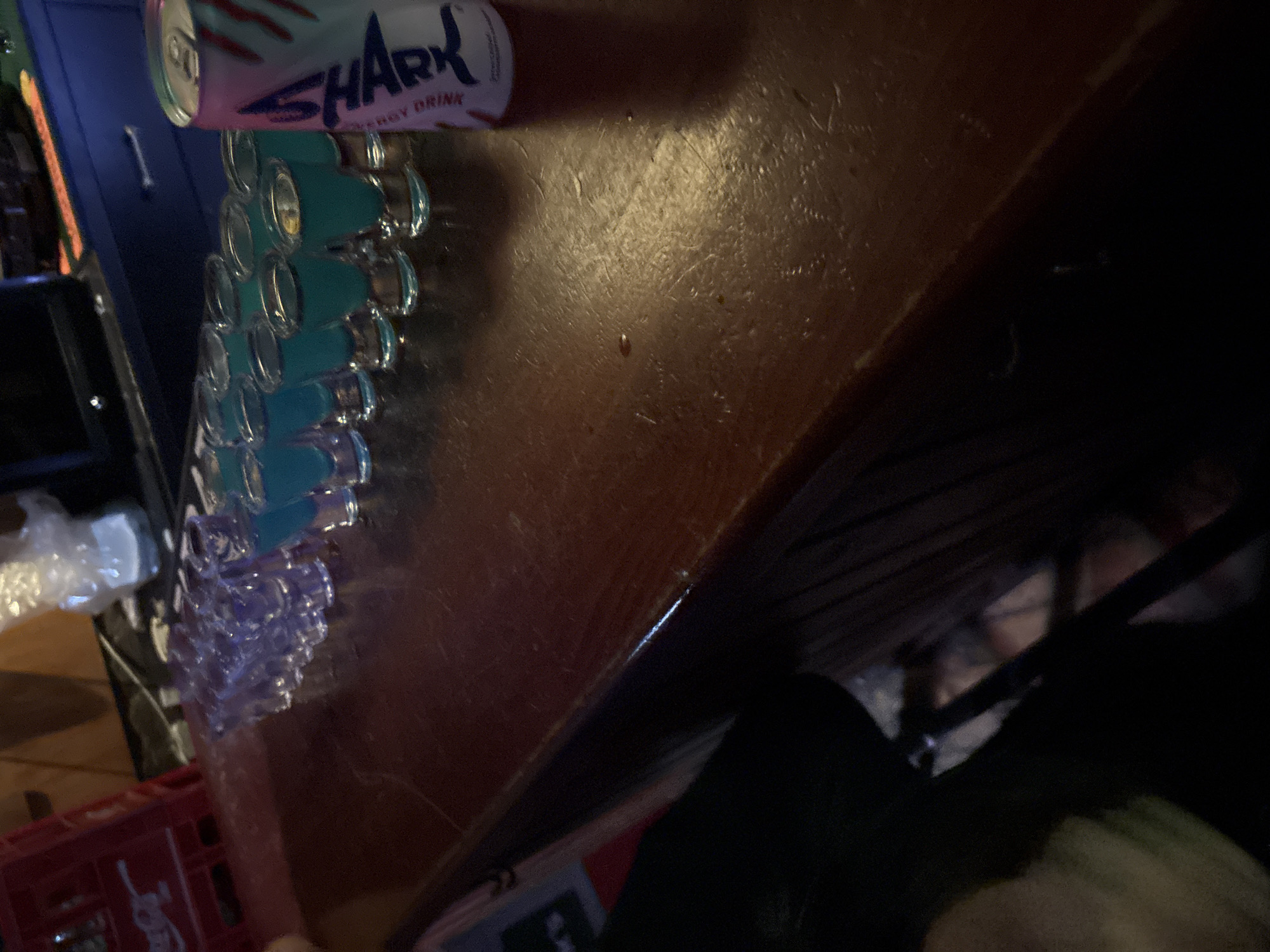 The image shows a bar setting, likely in Plovdiv, Bulgaria, given the request. A can of "Shark Energy Drink" sits on a wooden bar surface. Rows of shot glasses, some filled with a turquoise liquid and others clear, line the bar. A red plastic crate is visible in the lower left corner. A person is partially visible beneath the bar, obscured by shadows.