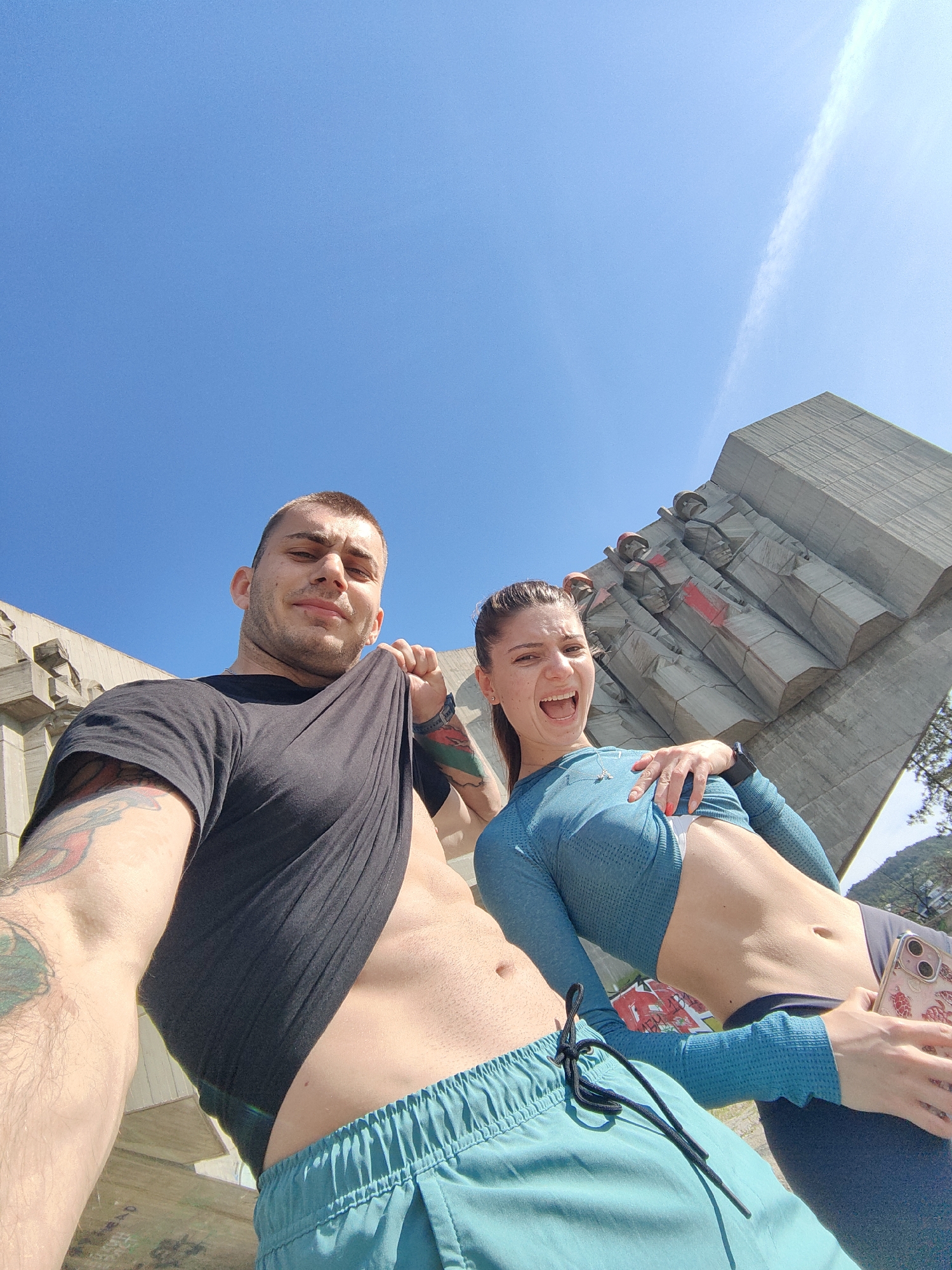 A low-angle shot shows a man and a woman posing in front of the Monument of the Bulgarian-Soviet Friendship in Varna, Bulgaria. The man, on the left, is Caucasian with a shaved head. He is pulling up his black t-shirt to expose his abdomen. Tattoos are visible on his left upper arm and left shoulder. He wears blue athletic shorts. The woman, on the right, is also Caucasian. She has her dark hair pulled back in a ponytail, is wearing a blue long-sleeved crop top, dark leggings, and holding a cell phone. She has her hand on her stomach and is smiling with her mouth open. The Monument of the Bulgarian-Soviet Friendship is in the background, consisting of large concrete figures with red accents. The sky above is clear and blue.