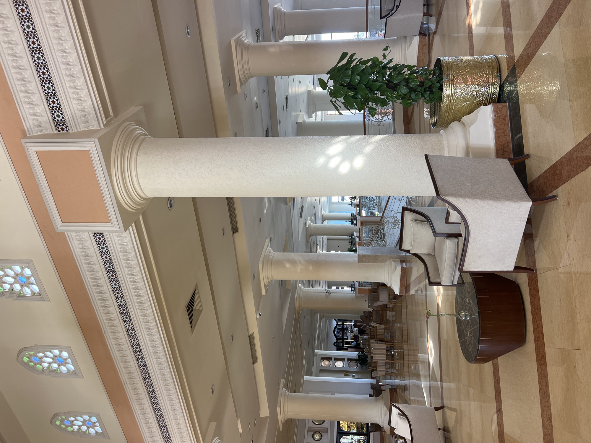 This image depicts the interior of a spacious lobby, likely in a hotel or resort, in Sharm El Sheikh, Egypt. The architecture features large, ornate columns, high ceilings, and decorative moldings. The floor is made of polished marble with inlaid patterns. Seating areas with plush armchairs and coffee tables are scattered throughout the lobby, suggesting a comfortable and welcoming atmosphere. In the background, dining tables and chairs are visible, hinting at a restaurant or dining area. Sunlight streams in from large windows, creating a bright and airy ambiance. The overall impression is one of elegance and luxury.