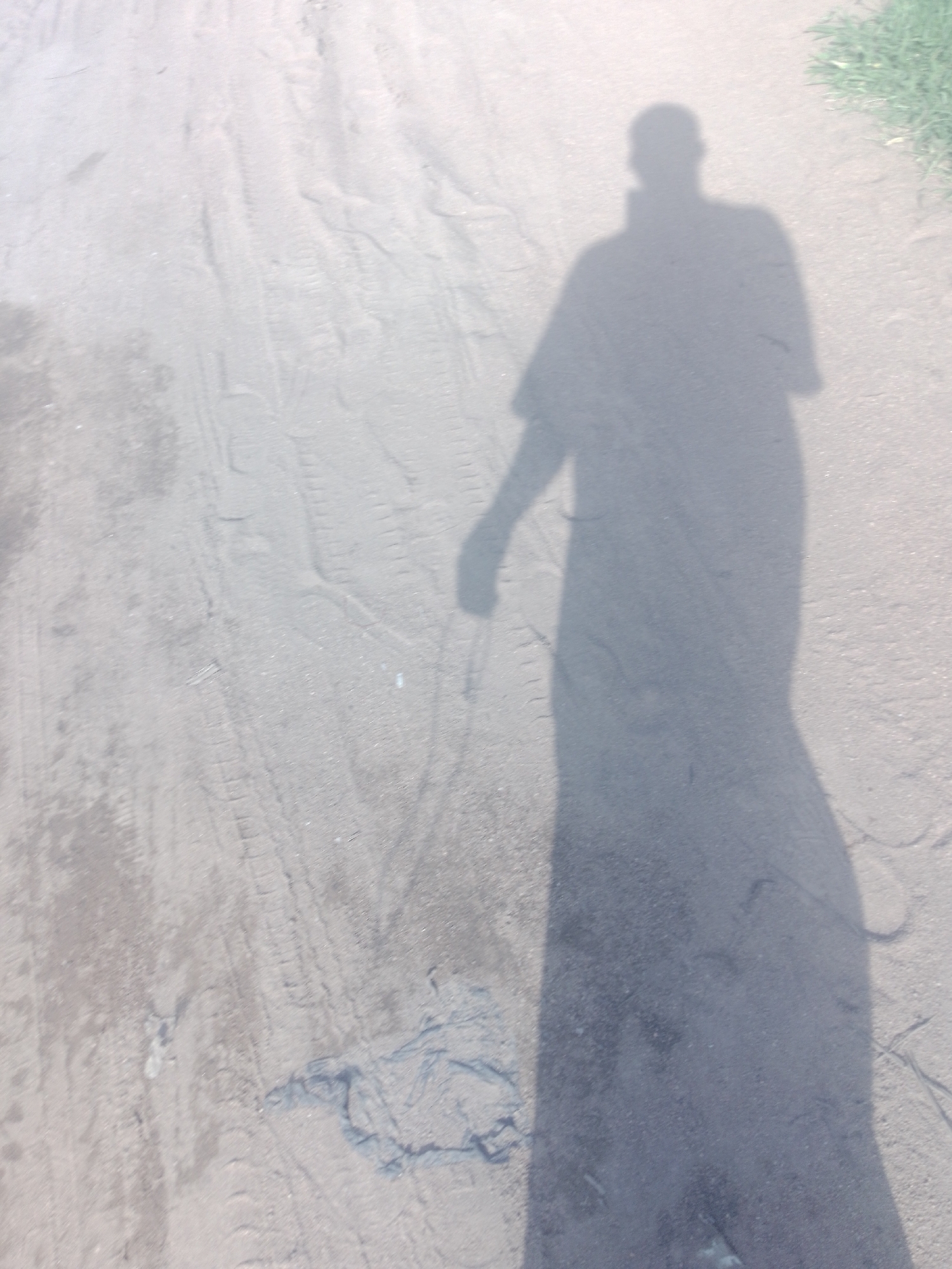The image shows a shadow of a person cast on a dirt road. The shadow is long and extends across the road, indicating a strong light source, likely the sun, from behind the person. The road surface appears to be rough and uneven, with tire tracks visible in the dirt. To the right of the shadow, a patch of green grass is visible, suggesting an outdoor setting. The overall lighting of the image is bright, with a slight overexposure, which contributes to the washed-out appearance of the scene. The location is identified as the city of Jalingo, Nigeria, implying a possibly arid or semi-arid environment.