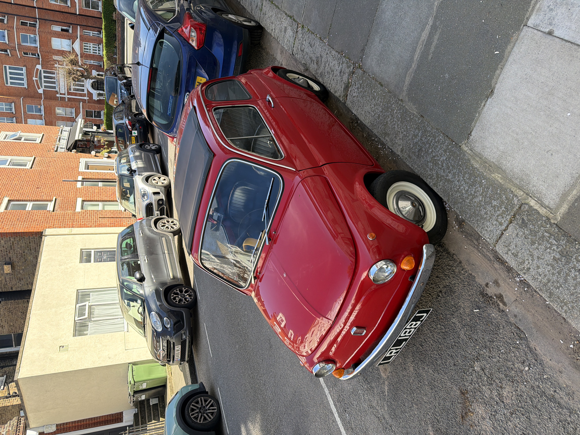 The image shows a street scene in London, United Kingdom. A red vintage car is parked alongside the curb, with its license plate reading "LRL 188J." Adjacent to the red car is a blue car, followed by other parked vehicles of varying colors and models. Buildings, mainly brick apartment buildings, line the opposite side of the street. The street appears to be in a residential area. The weather seems sunny, with shadows visible. A green recycling bin is visible in the lower left of the image.