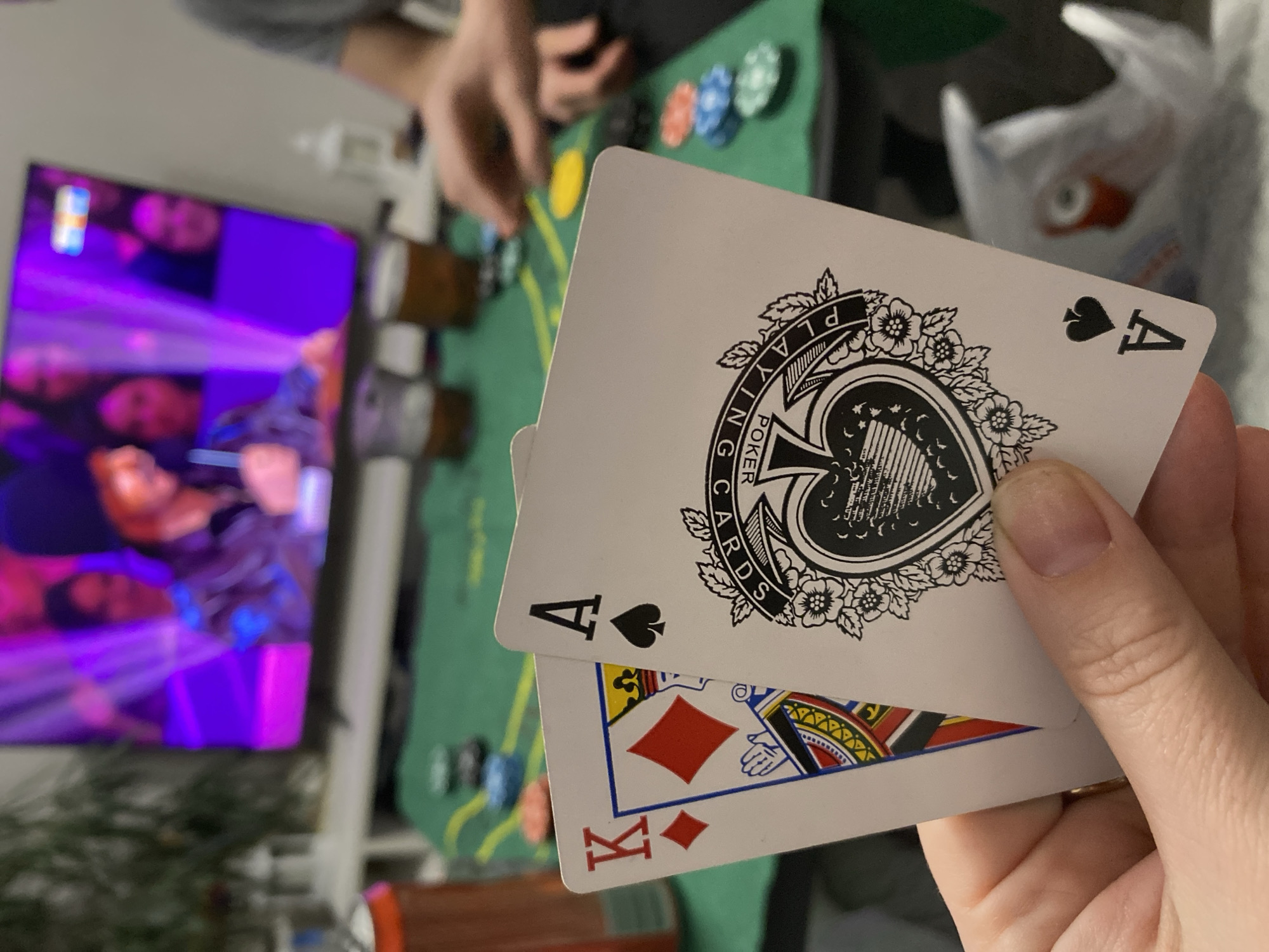 Here is a detailed description of the image:
**People:**
* A person is holding playing cards in their hand.
* Another person is visible in the background at the poker table.
* People are also visible on the television screen.
**Subjects/Objects:**
* Two playing cards are prominently displayed: the Ace of Spades and the King of Diamonds. The Ace of Spades has decorative artwork around the spade symbol.
* There is a green poker table with poker chips.
* A television screen shows people, with primarily purple and blue hues.
* A white plastic bag is visible in the background.
**Actions/Interactions:**
* The person holding the cards is likely participating in a poker game.
* Someone in the background is also likely participating in the game.
* The television is on, providing entertainment in the background.
**Environment:**
* The scene appears to be indoors, possibly a home.
* The lighting is somewhat dim.
* The background includes a TV and other miscellaneous items.
**City Location (Chaikovsky, Russia):**
* There is no explicit information in the image to definitively confirm it was taken in Chaikovsky, Russia. Any association with that city is based on external factors not present in the visual data.