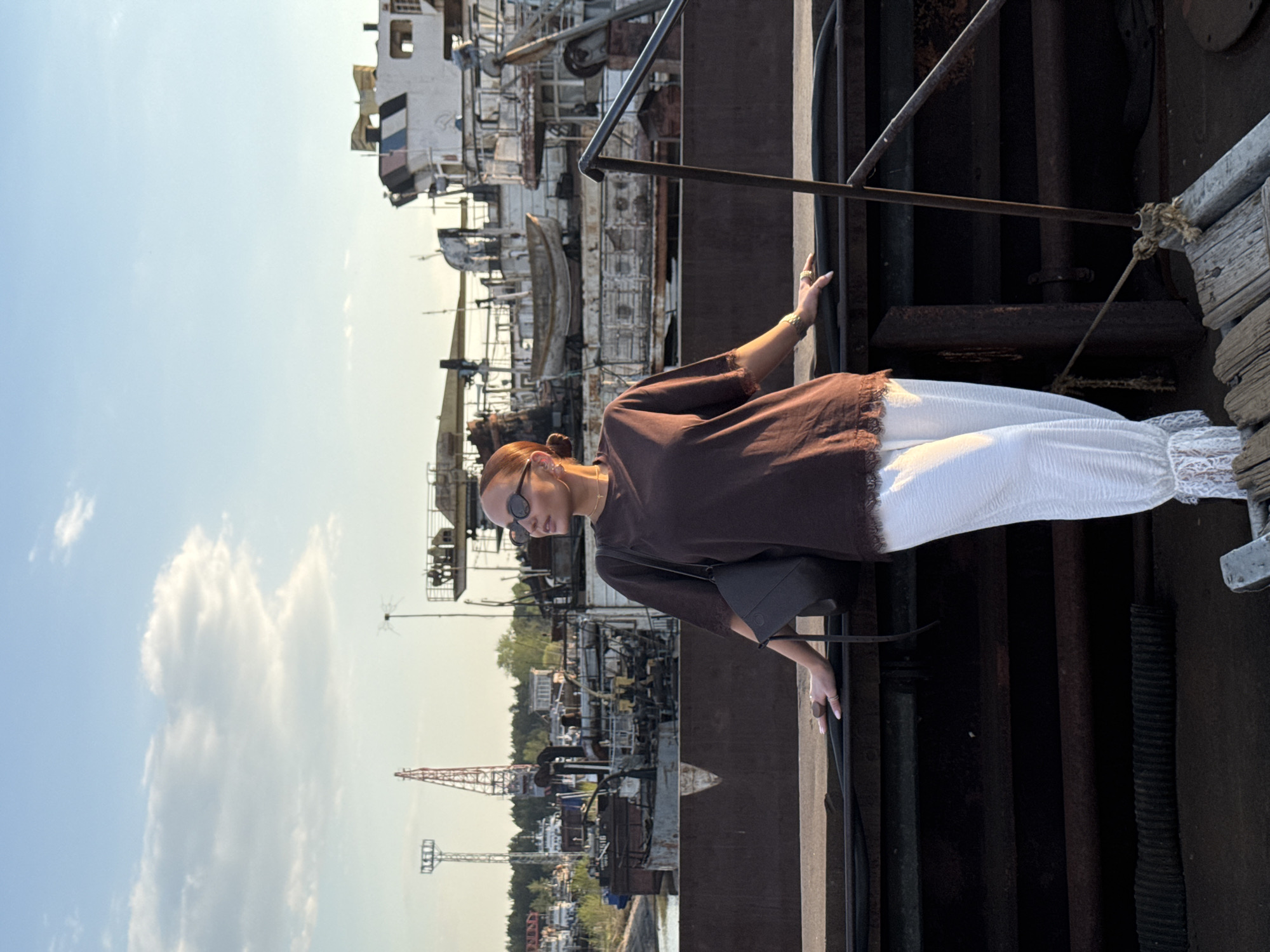 A full-body shot shows a woman with auburn hair styled in a bun, wearing sunglasses, a brown tunic with fringed sleeves, and white pants with lace trim, standing on a weathered metal structure. She is holding onto a metal railing. In the background, there are numerous metal constructions, possibly old ships, under a blue sky with light clouds. Cranes are visible in the distance. This is likely a dock or industrial area in Chaikovsky, Russia, based on the provided location information.