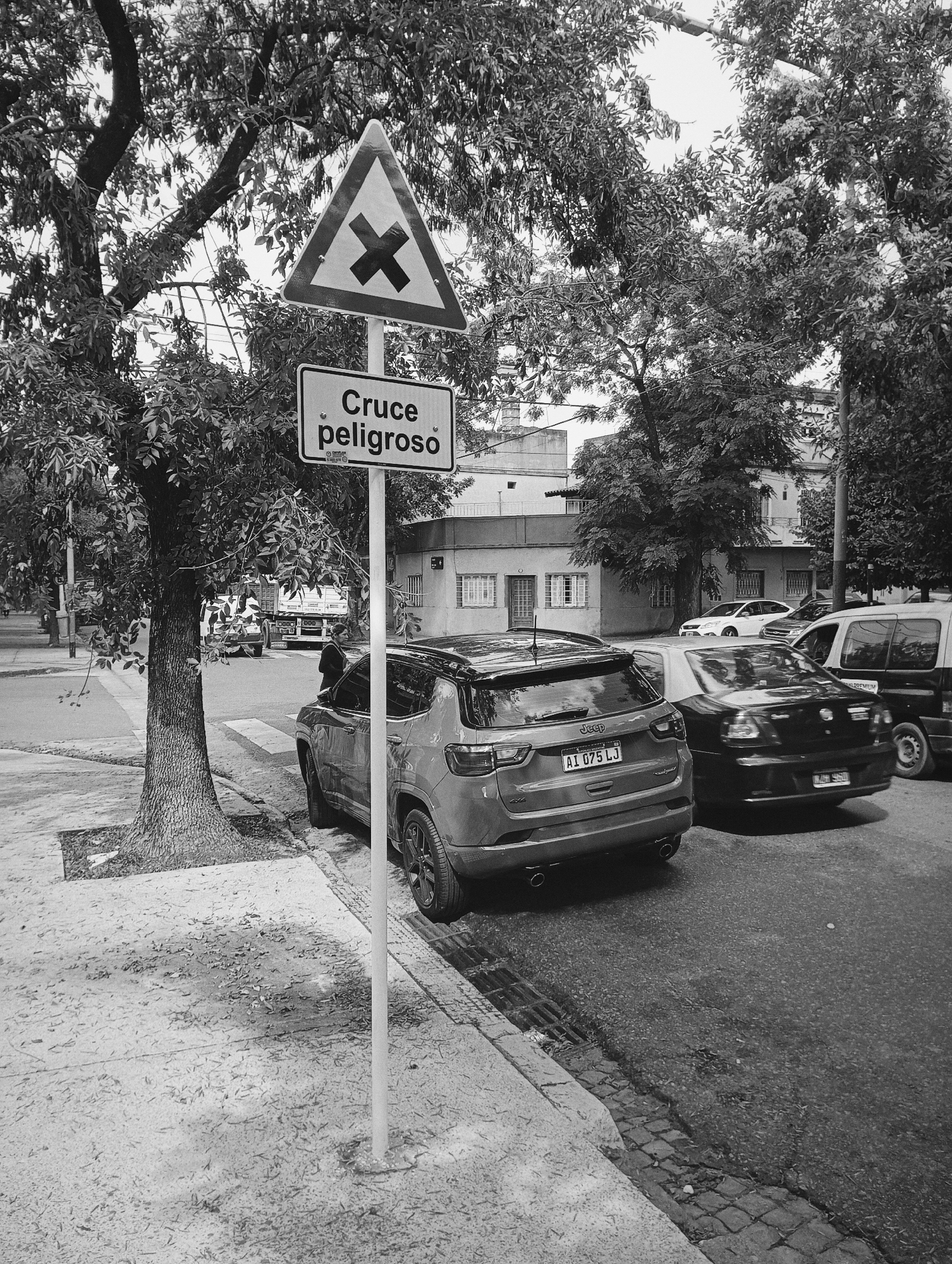 The image shows a street scene in Buenos Aires, Argentina. A traffic sign is visible in the foreground, indicating a dangerous crossing ("Cruce peligroso"). The sign consists of a triangular hazard symbol above a rectangular sign with the text "Cruce peligroso."
A Jeep Compass with license plate "AI 075 LJ" is parked next to the sign. To the right of the Jeep, a black car is partially visible, its license plate partially obscured. Further down the street are other cars, including a white sedan and a taxi. In the background, a truck is visible on the crossing.
The scene is typical of an urban residential street with trees lining the sidewalk. The sidewalk is paved and slightly worn, and leaves litter its surface. The buildings are two-story residential units. The photograph is black and white. The overall scene suggests a typical urban environment in Buenos Aires.